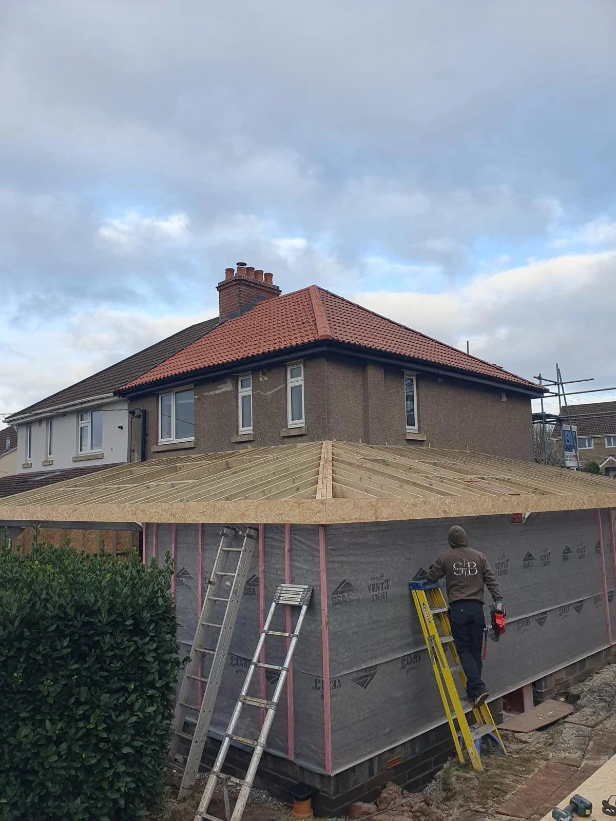 A wooden framework of a building under construction with scaffolding and safety fencing, and a partially finished roof with tiles.