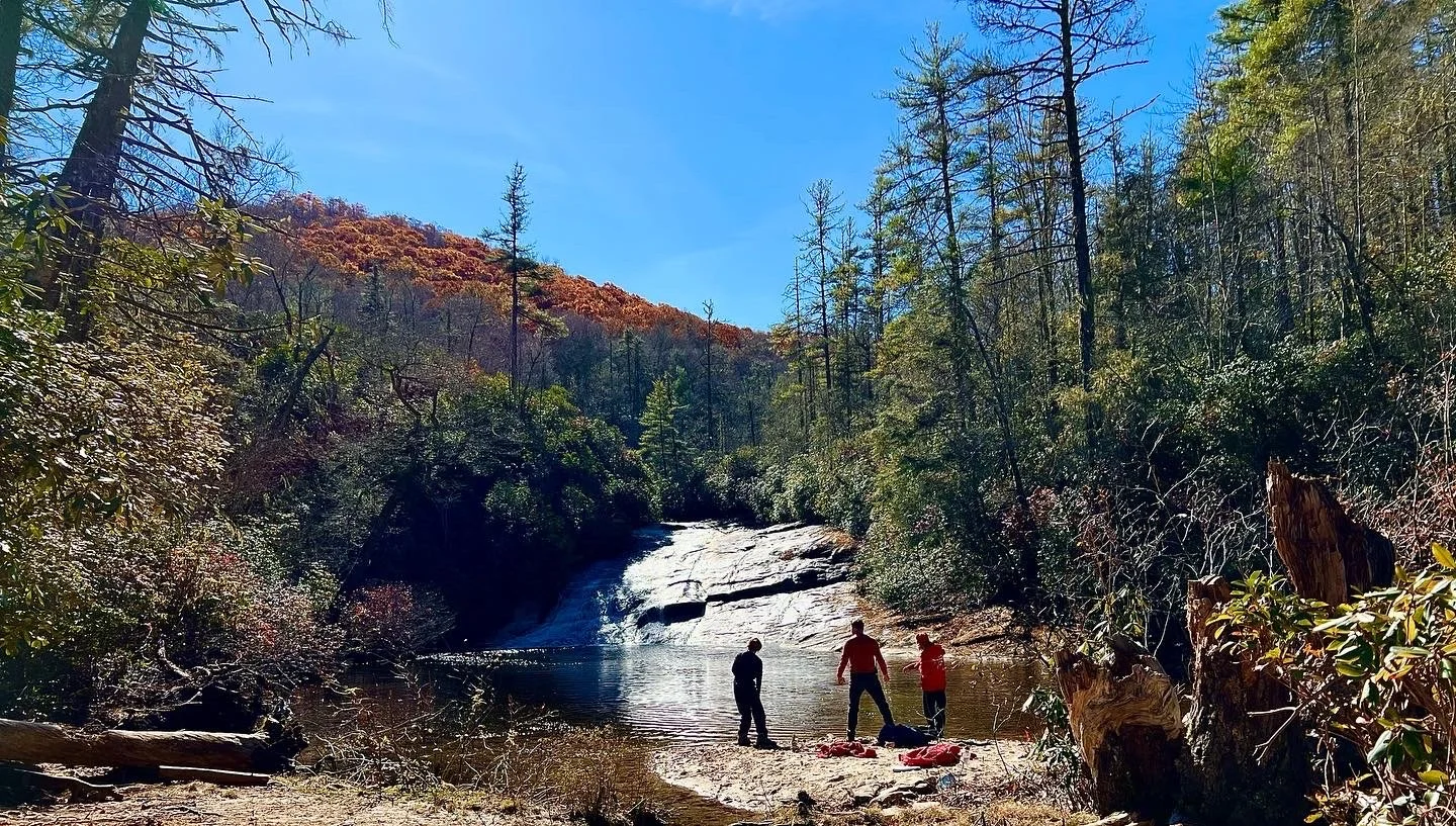 Three people standing near a small river or pond in a forested area with trees and a rocky waterfall in the background under a bright blue sky.
