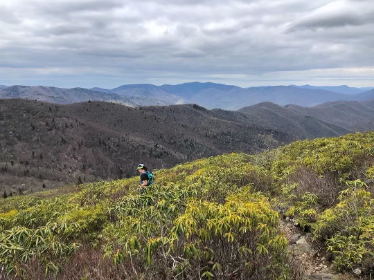 Hiker in a green, hilly landscape with mountain ranges in the background under cloudy sky.