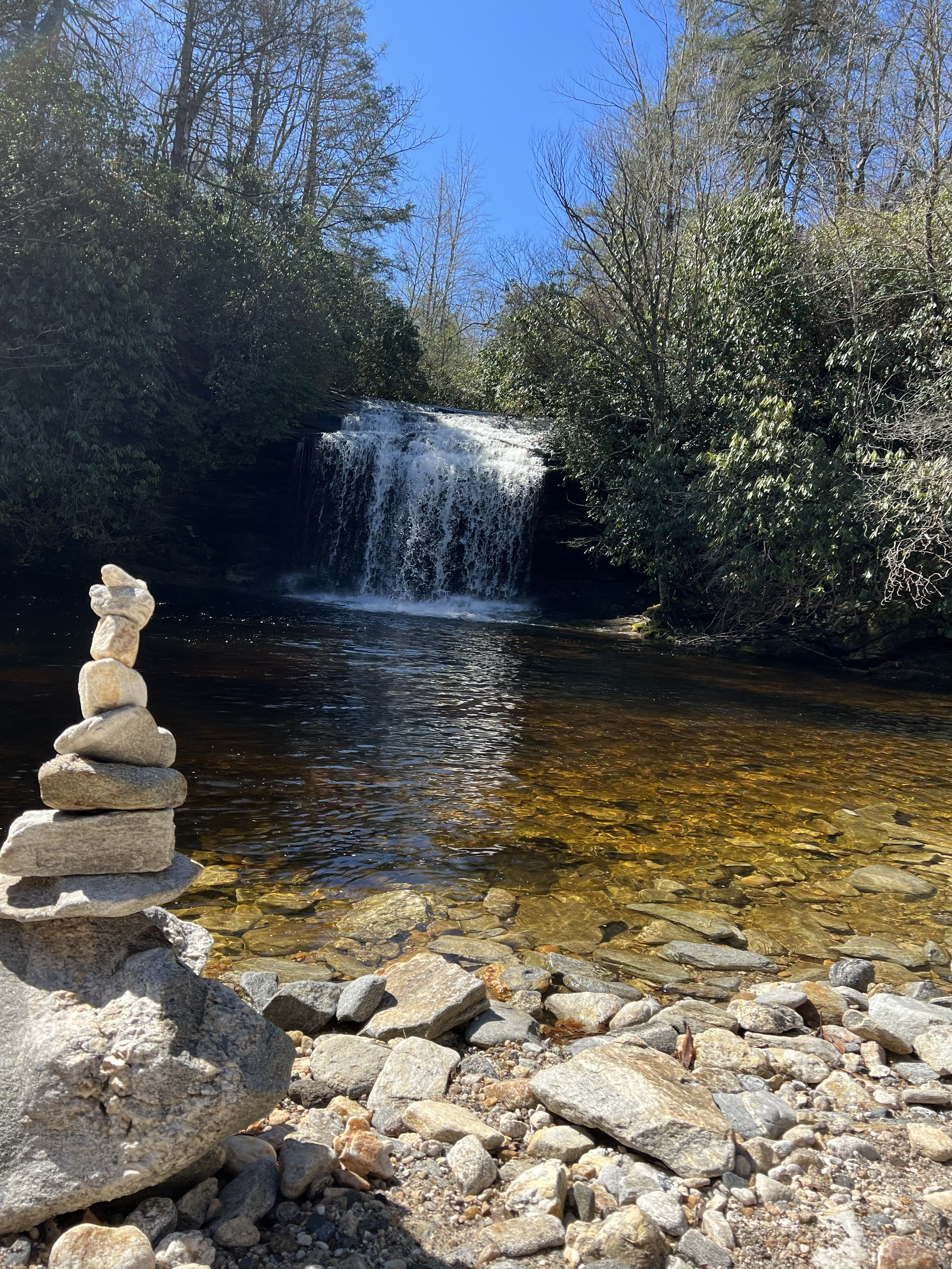 A small waterfall flowing into a river surrounded by trees and rocks, with a stone cairn in the foreground.