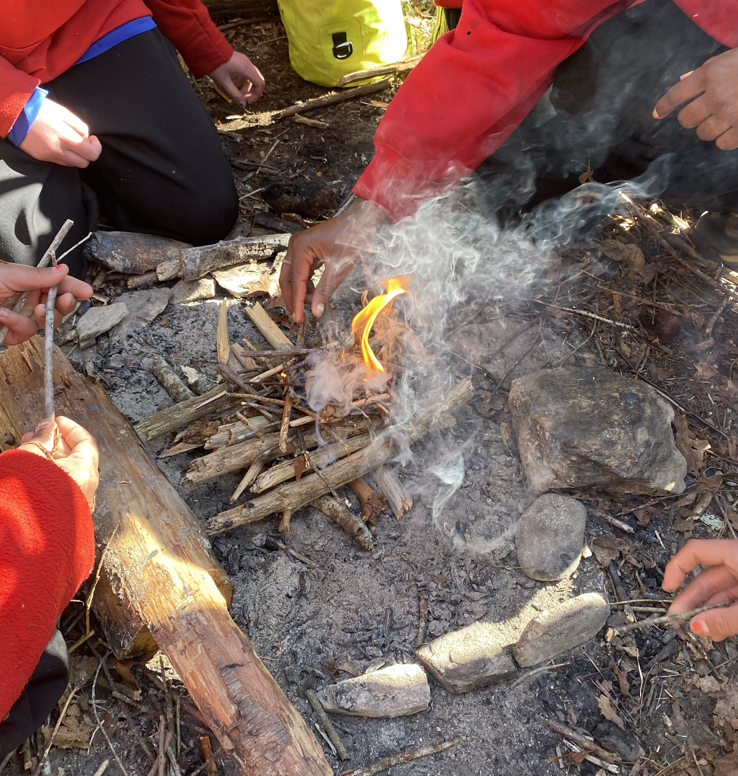 A group of people building a small campfire with sticks and kindling in a forested area, with some hands holding matches or small sticks near the fire.