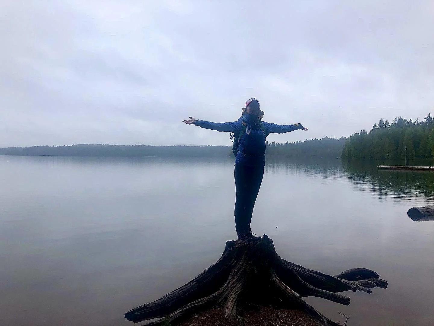 Person standing with arms outstretched on a fallen log at the edge of a calm lake, surrounded by trees under an overcast sky.