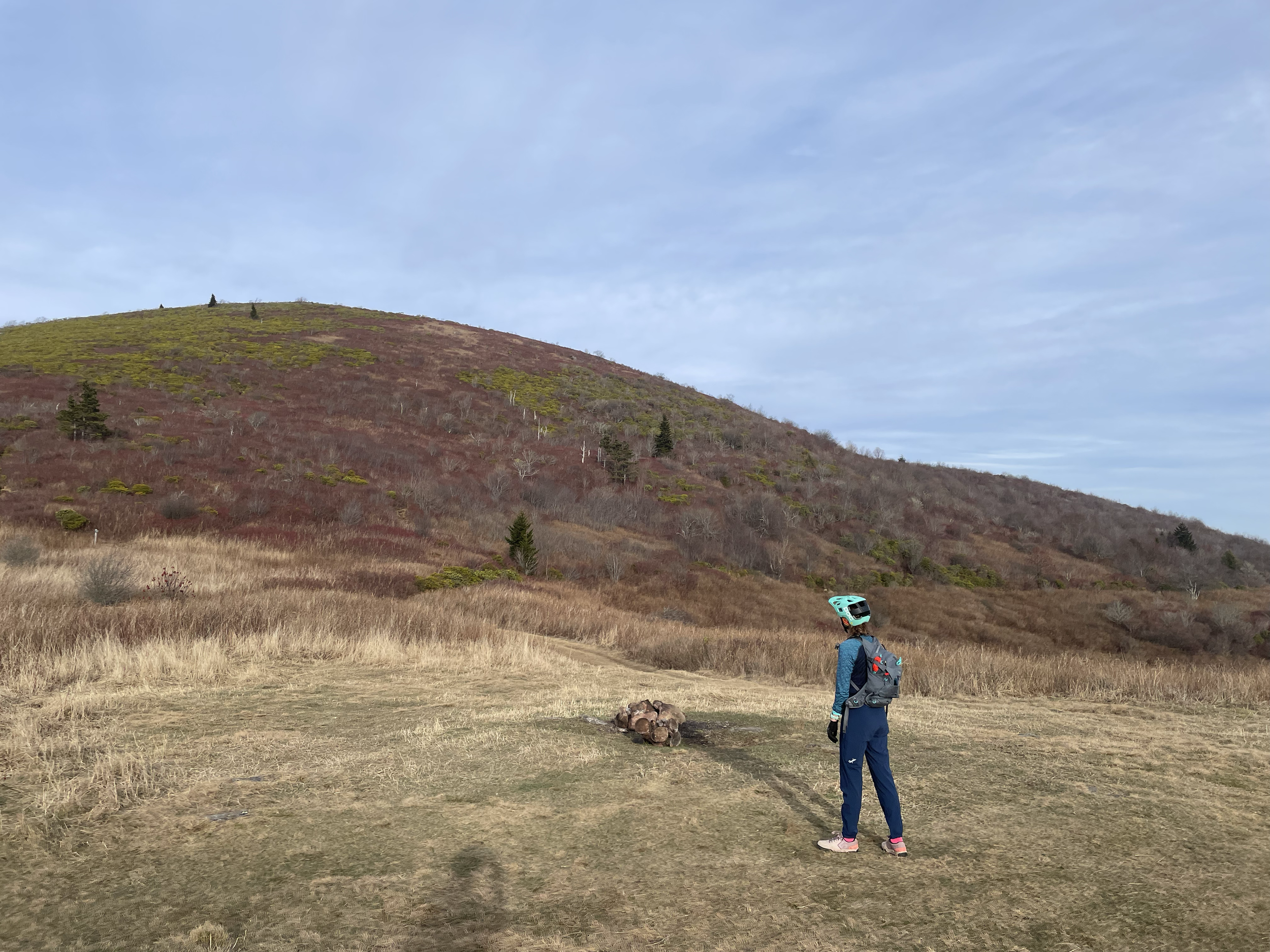 Person in cycling gear, including a helmet and backpack, standing on a dirt trail in a hilly, grassy landscape during daytime, with a mountain covered in sparse vegetation in the background.