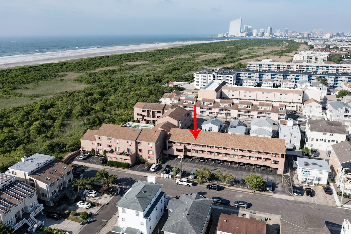 Aerial view of a coastal town with a beach, a green nature area, and high-rise buildings in the distance, featuring a large residential building with a red arrow pointing to it.