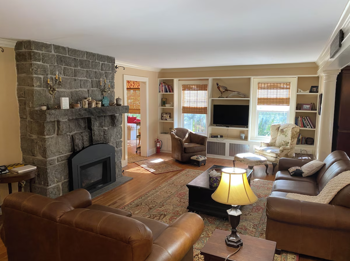 Living room with stone fireplace, brown leather sofas, a floral armchair, a television on built-in shelves, and a patterned area rug.