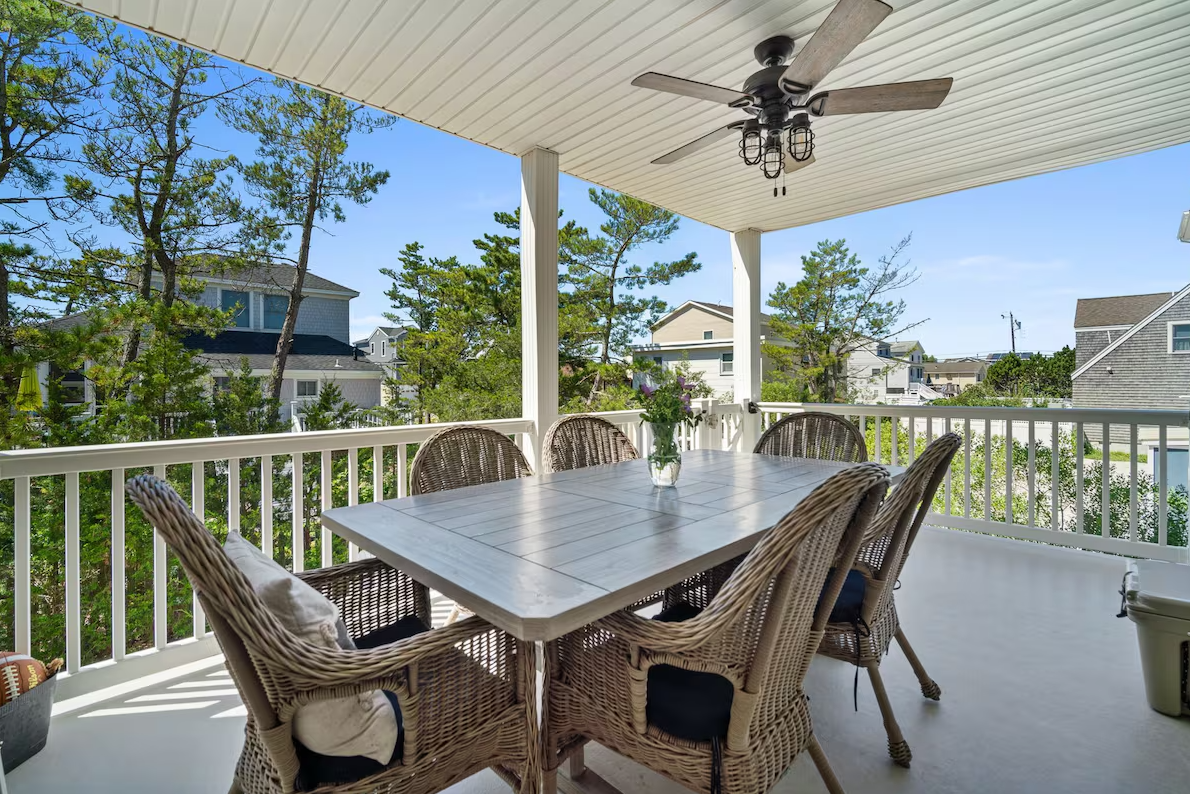 A covered balcony with a white wooden table, six wicker chairs with cushions, a vase with flowers, and a ceiling fan with light fixtures, overlooking trees and neighboring houses.