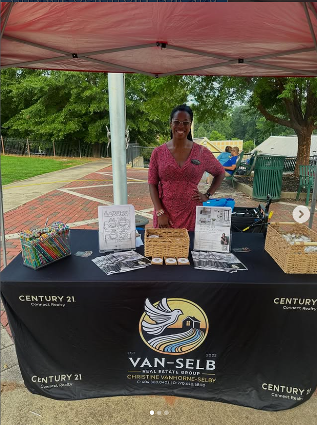 Woman standing at a table with real estate branding, under a pink canopy outdoors with trees and a park background.