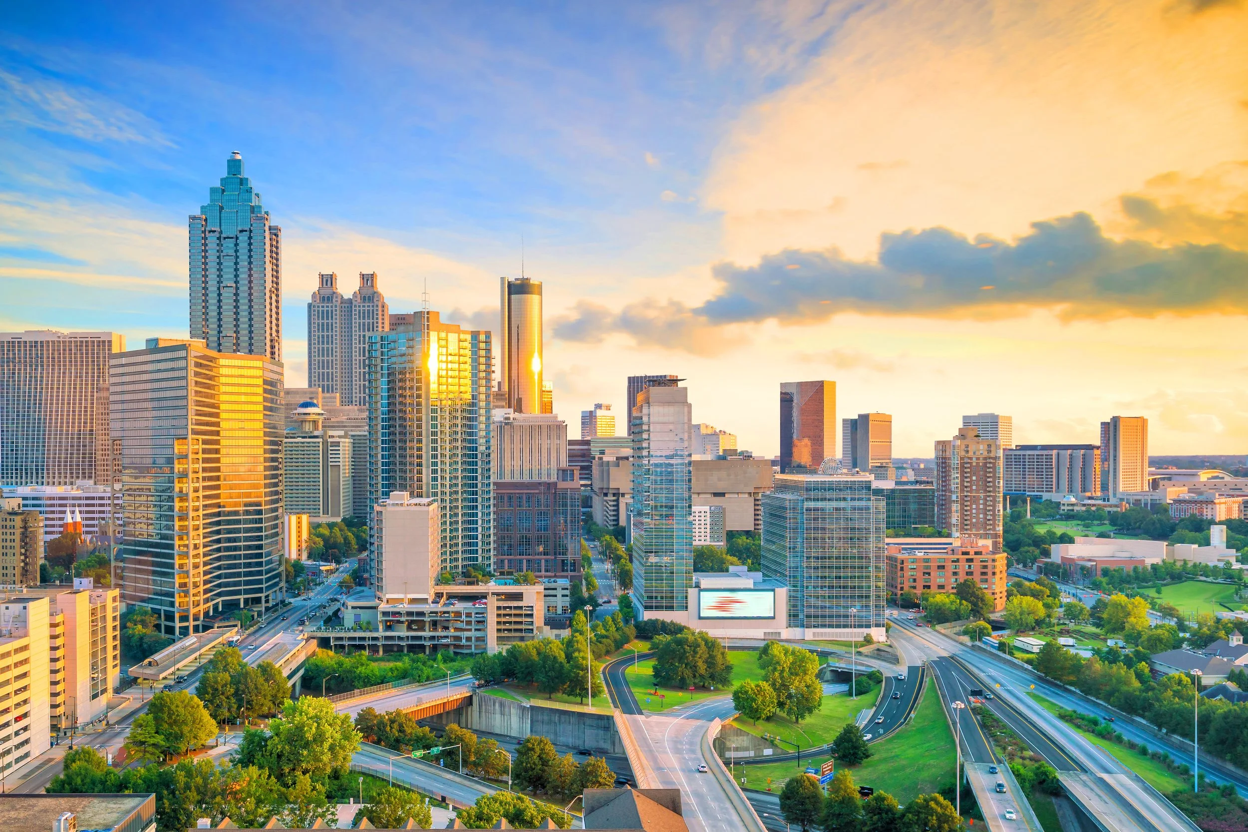 Skyline of Atlanta, Georgia during sunset with tall modern skyscrapers, roads, and green parks in the foreground.