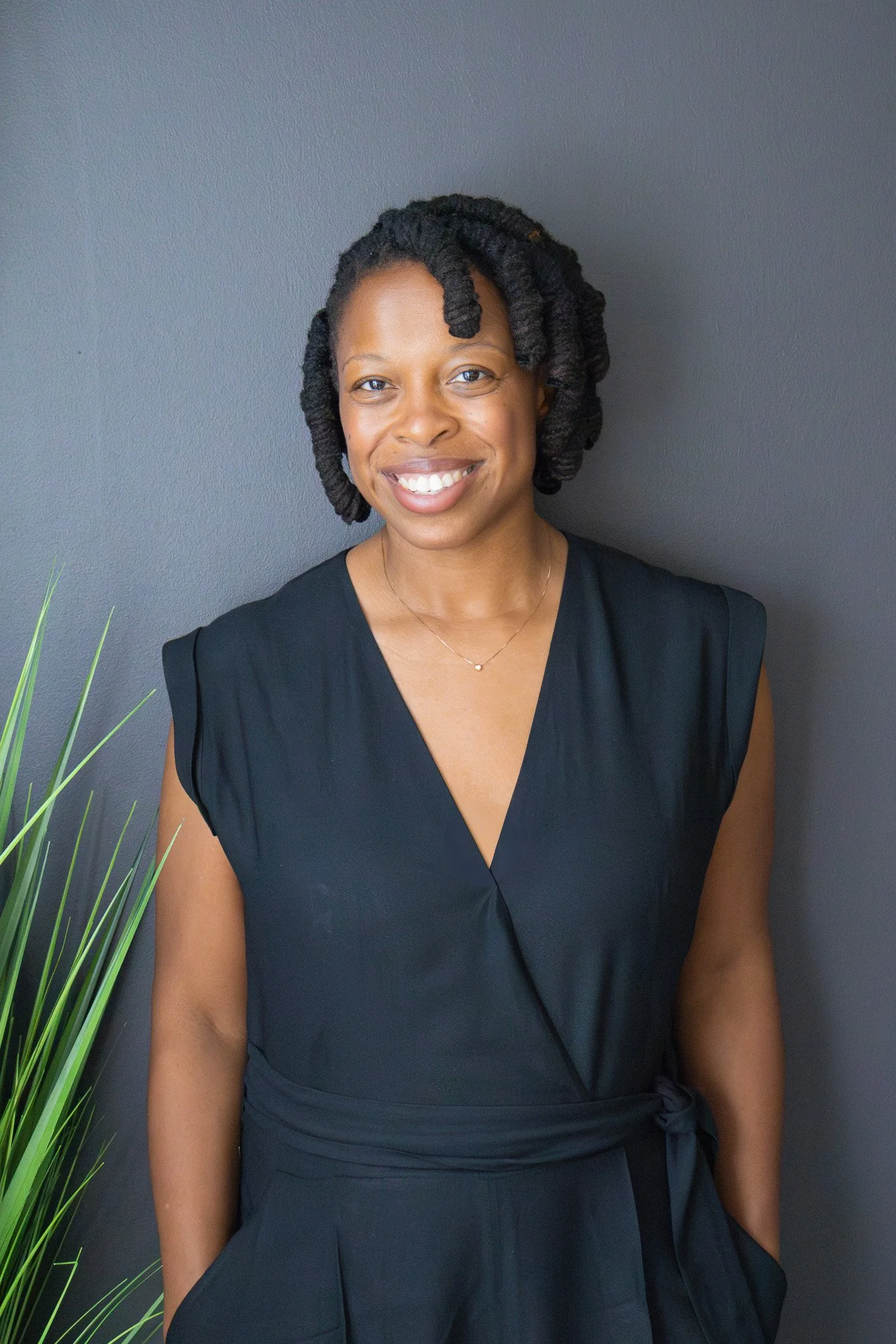 A smiling Black woman with styled natural hair, wearing a sleeveless black wrap dress, standing against a dark gray wall with some green plant leaves visible on the left.