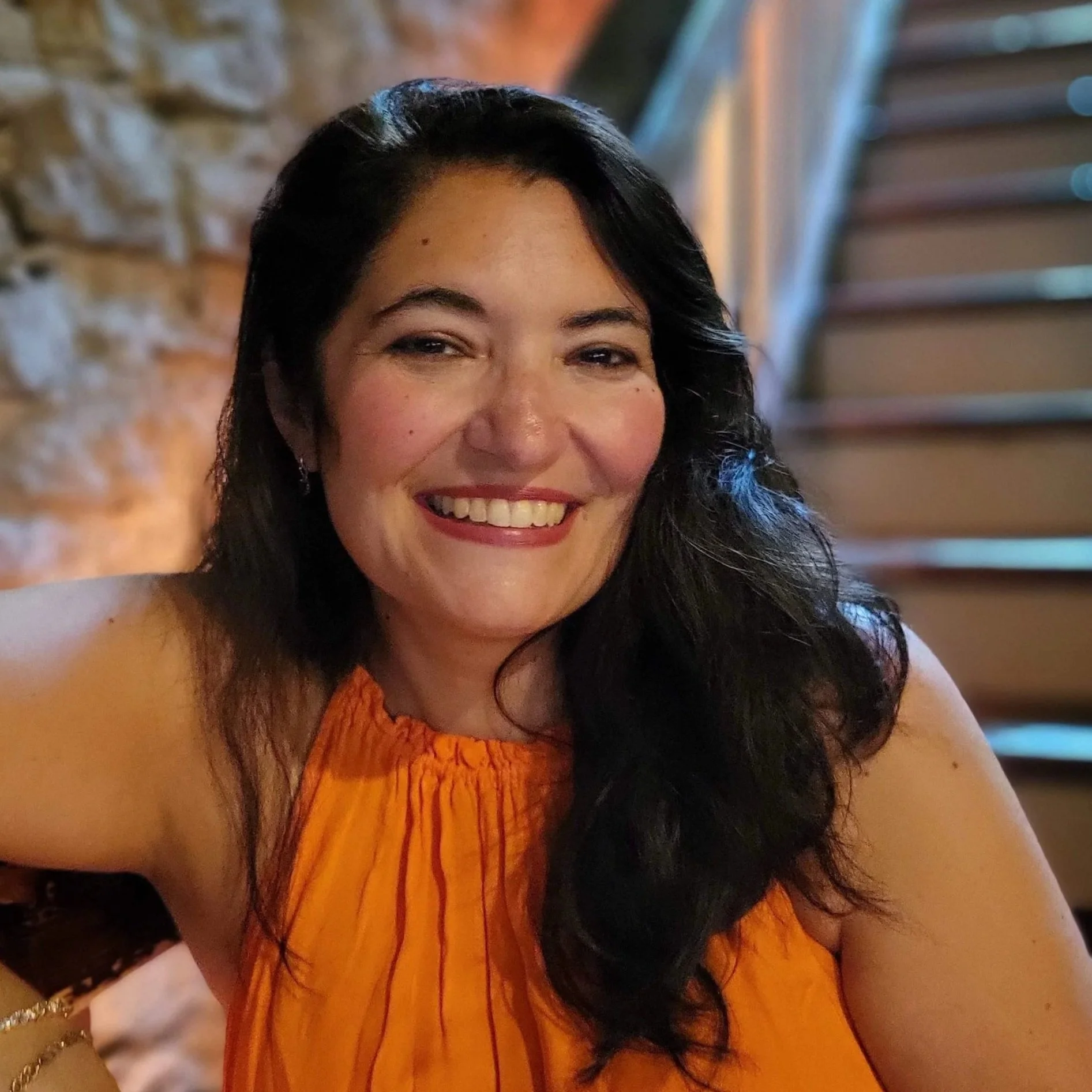 Smiling woman with dark hair in an orange sleeveless top, sitting against a stone wall near a staircase.