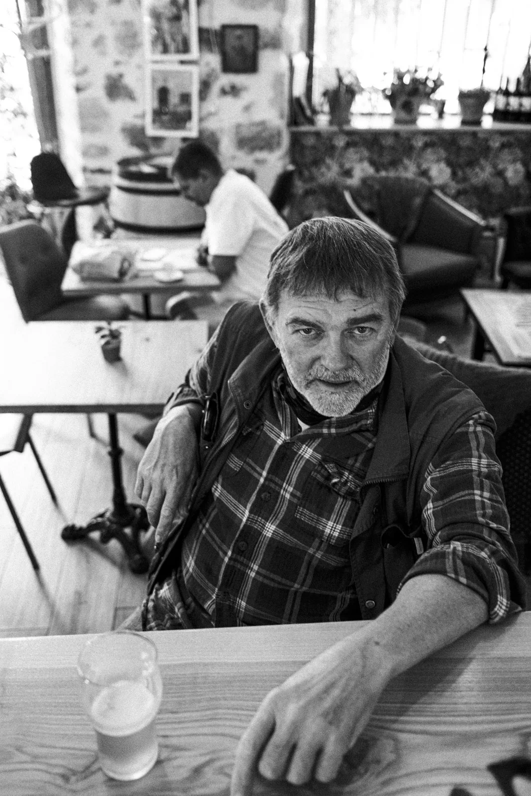 A black and white photo of a man with a beard and gray hair sitting at a table in a cafe, looking at the camera. In the background, there are two other people sitting at a table, one eating. The cafe has stone walls, framed pictures, and windows with