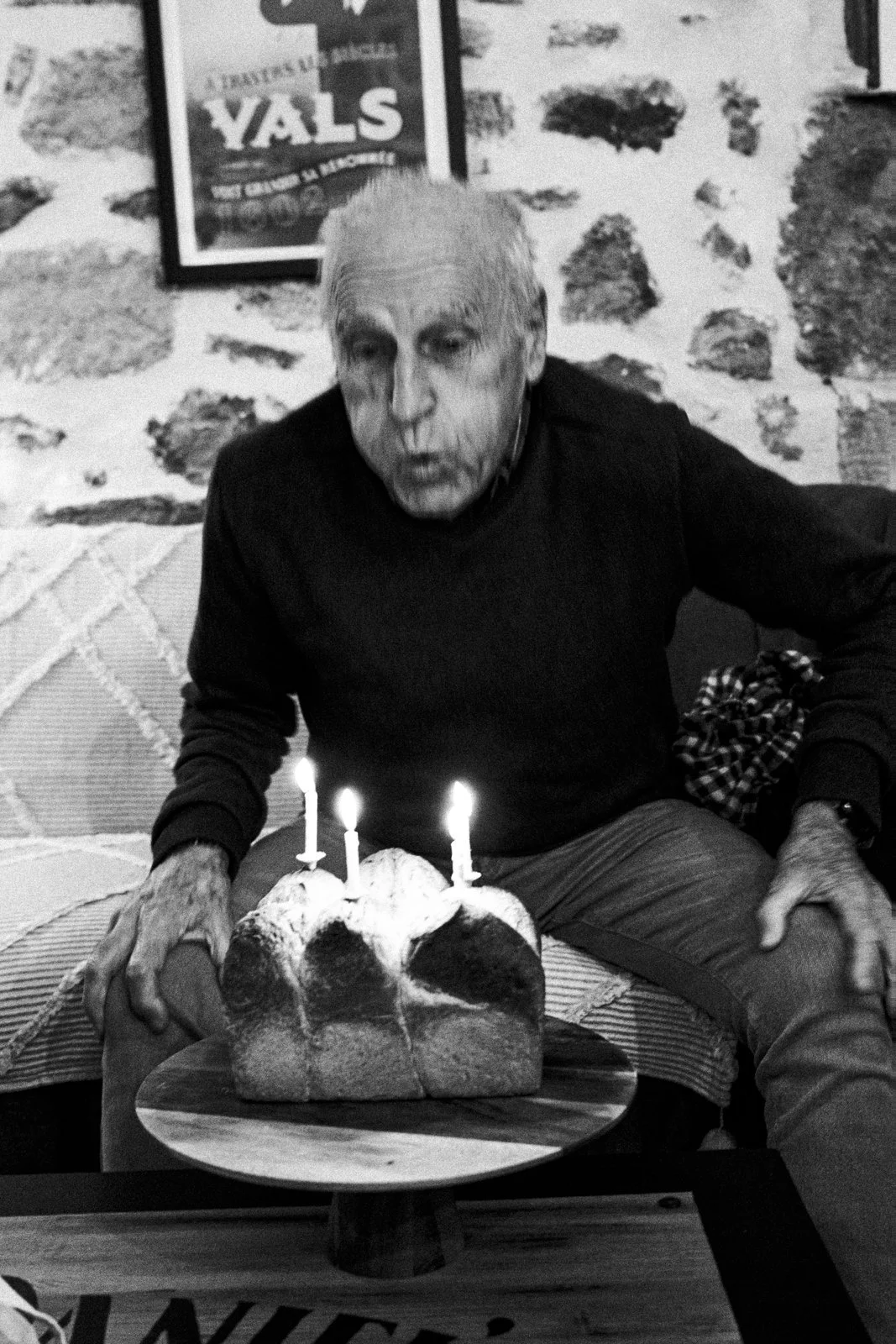 An elderly man sitting at a table with a cake that has three lit candles on top, blowing out the candles.