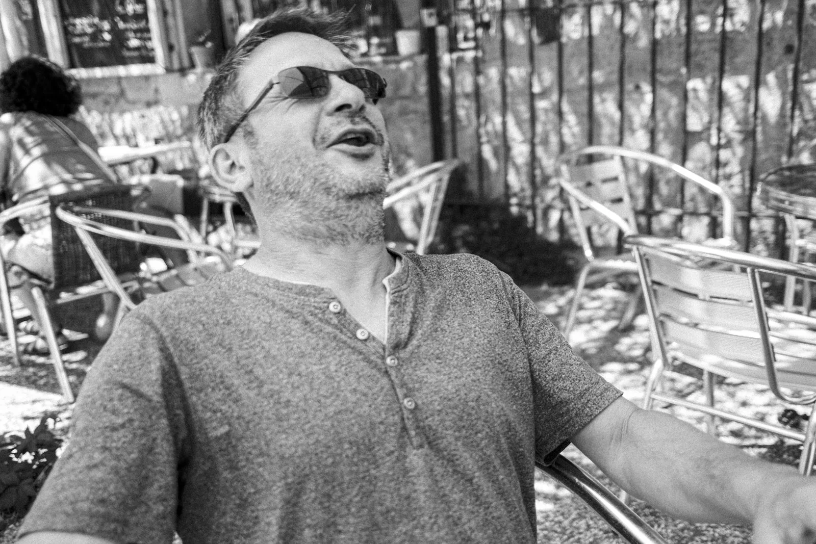 A man wearing sunglasses and a textured henley shirt sitting outdoors on a patio with empty chairs around him.