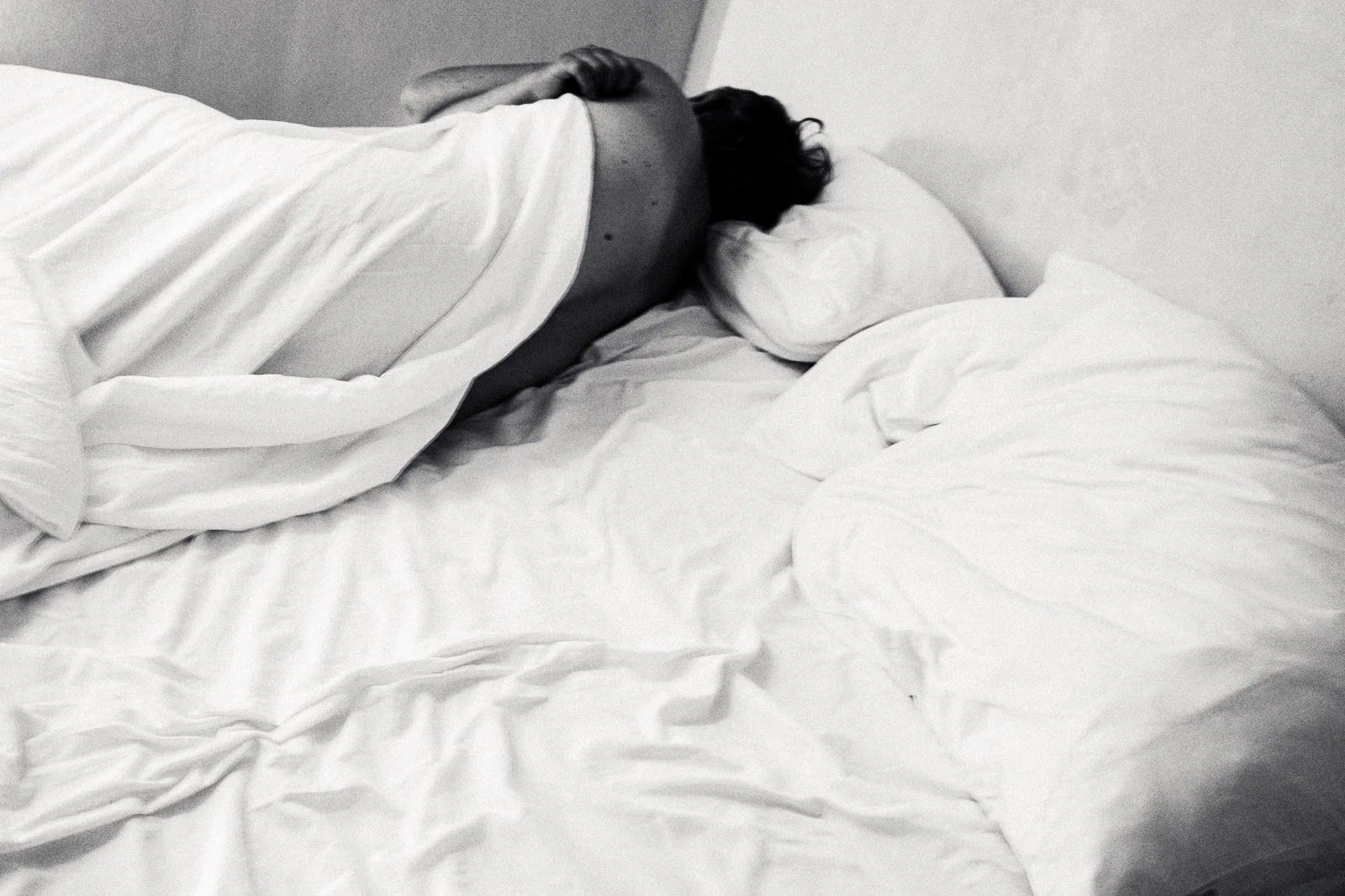 A person sleeping on a bed with white sheets and pillows, resting on their side with arms under their head, in black and white photo.