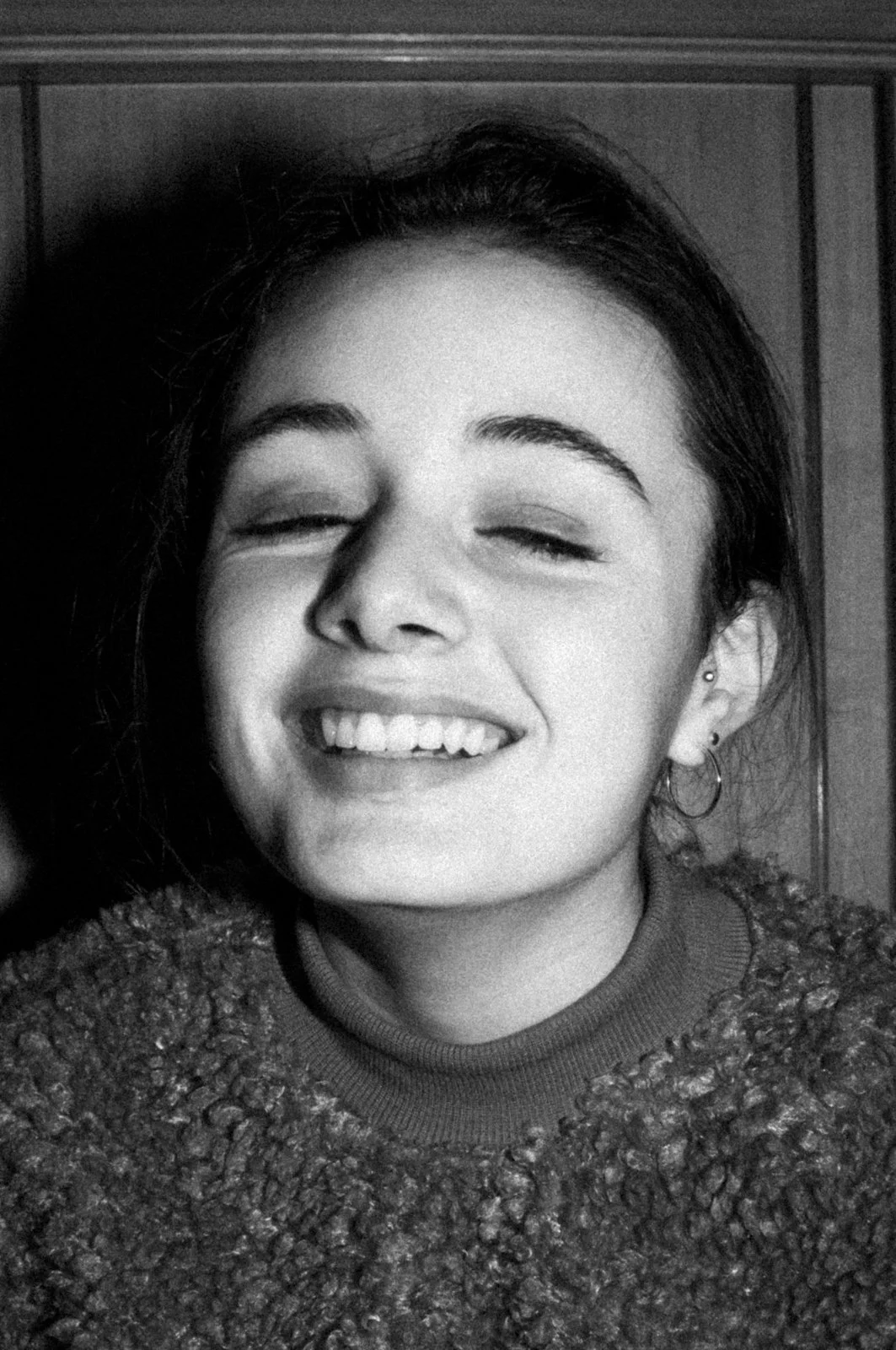 Black and white photo of a young woman smiling with eyes closed, showing her teeth, wearing earrings and a textured sweater.