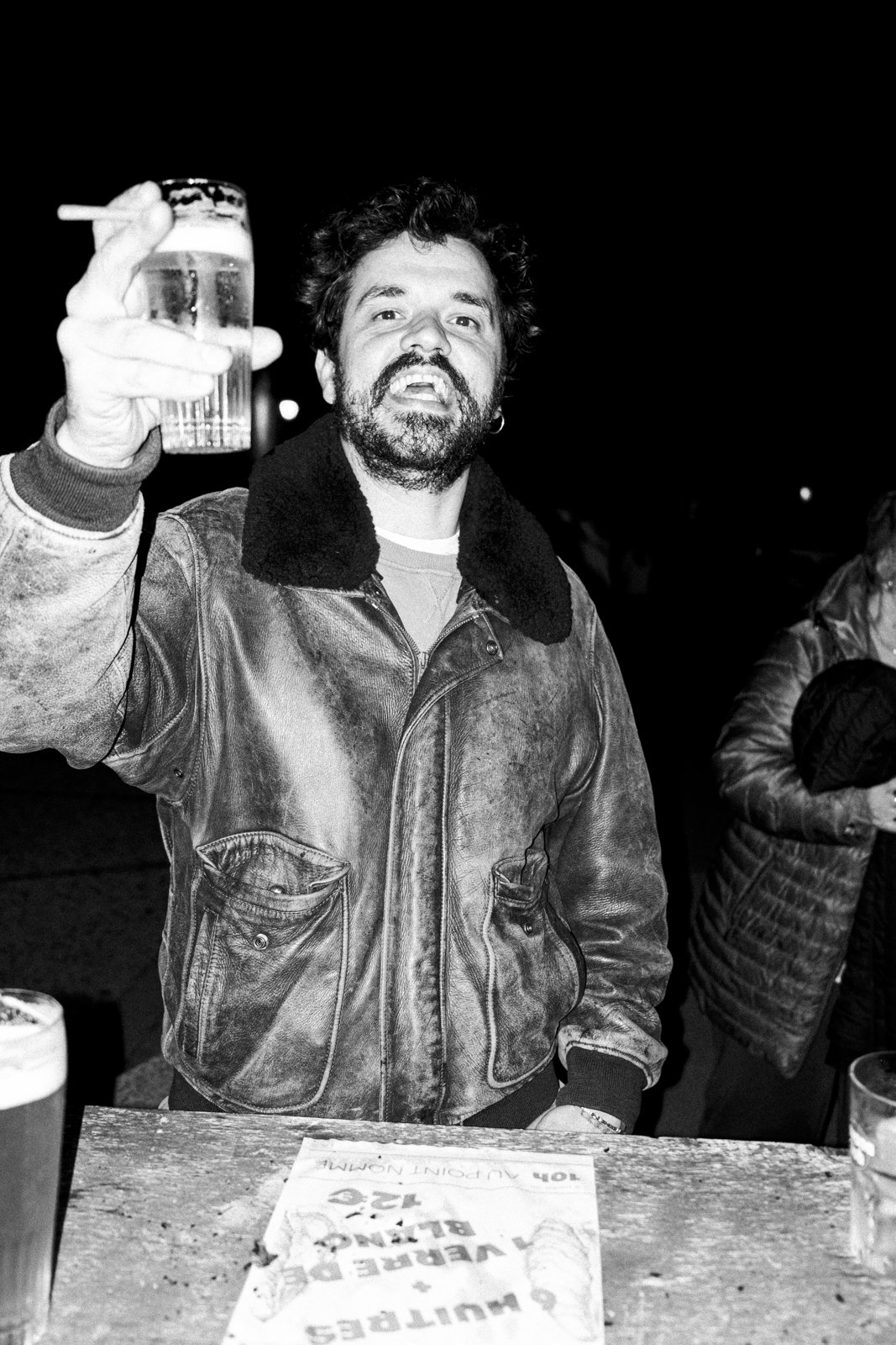 Man with thick beard and curly hair holding a drink and cigarette in a bar at night, wearing a leather jacket with a fur collar.