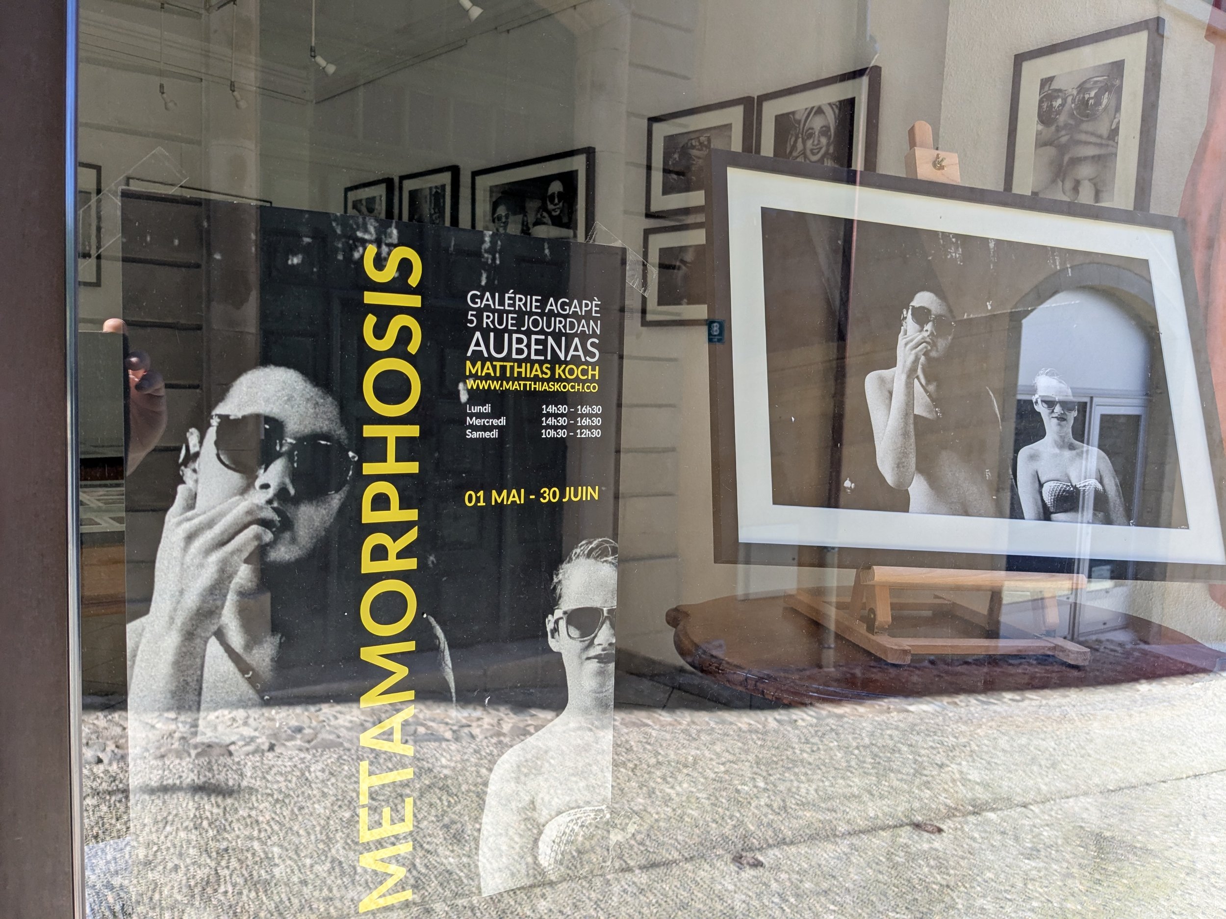 Gallery window display featuring a black and white poster for 'Matthias Koch Metamorphosis' art exhibition, with an acrylic display of two black and white photographs of women in sunglasses, one holding a cigarette, inside the gallery.