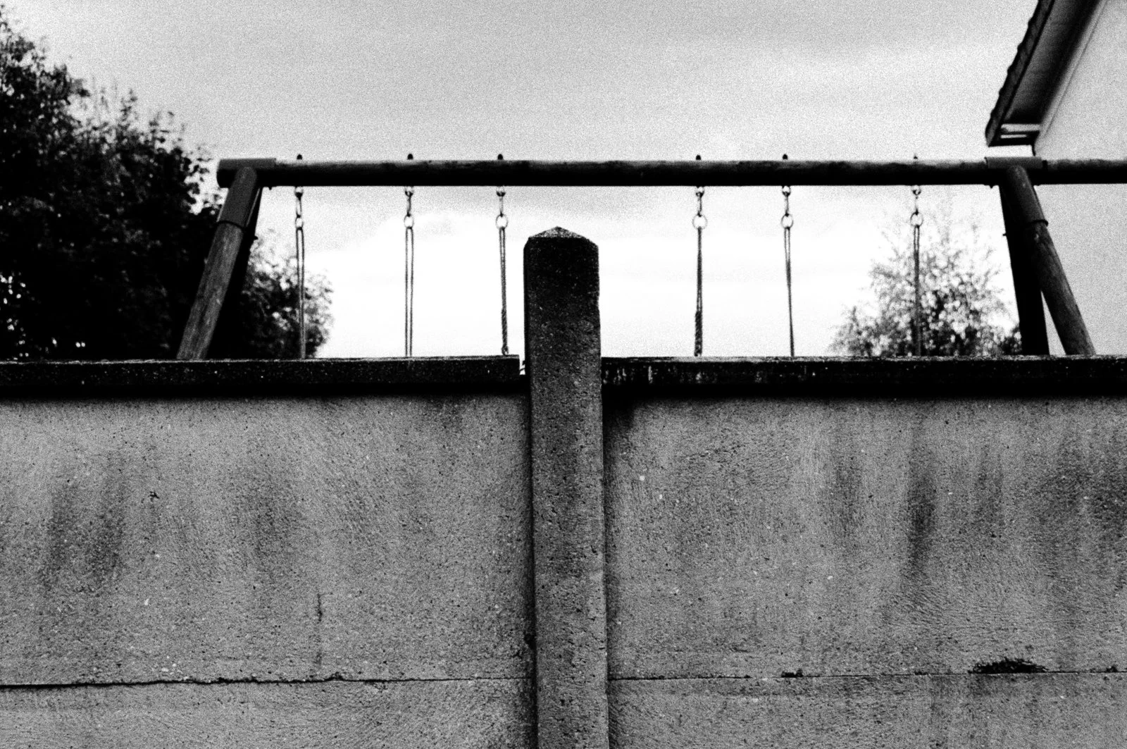 Black and white photo of a fence on top of a concrete wall, with trees and sky in the background.