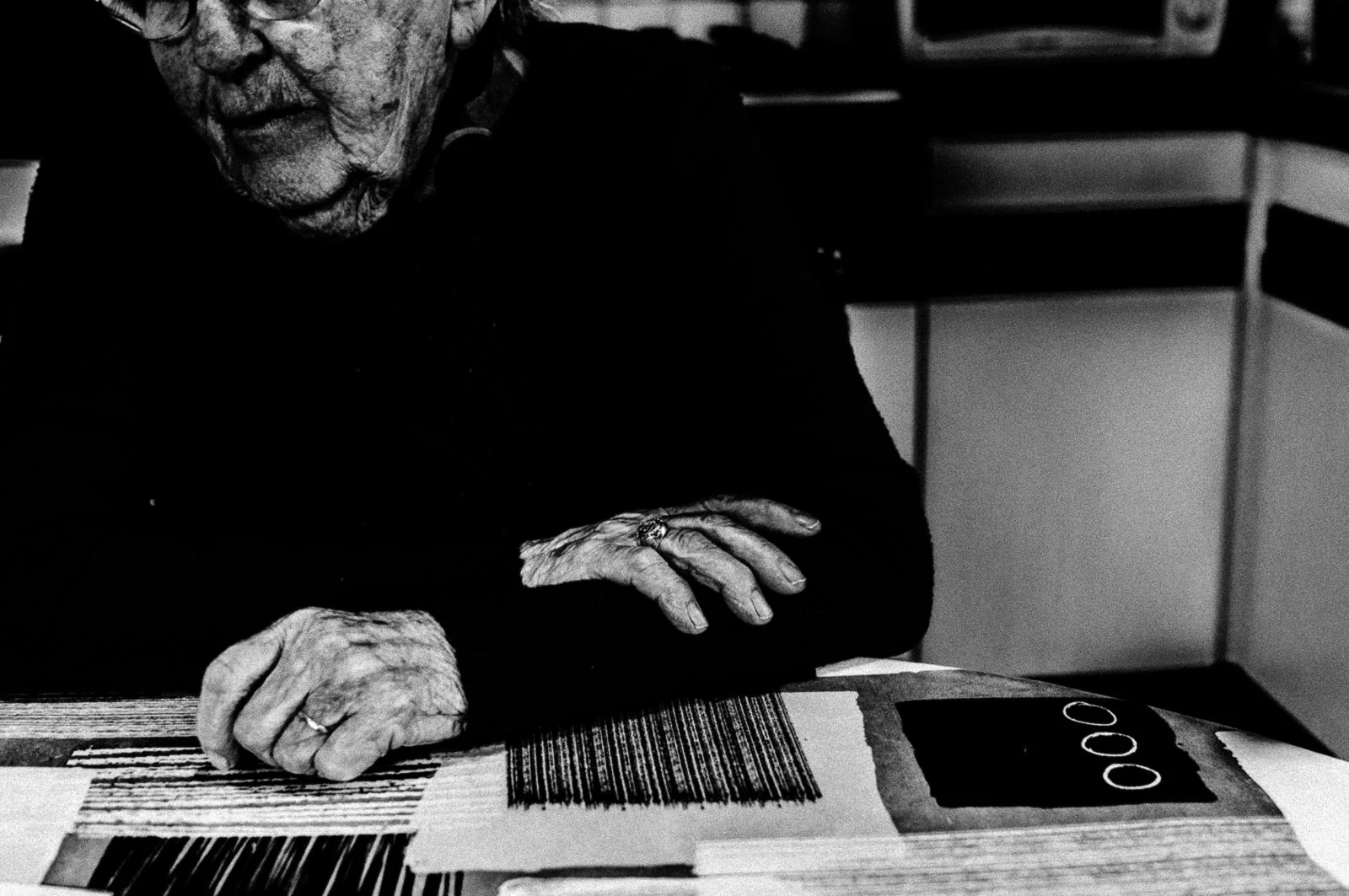 Close-up of an elderly woman's hands resting on a wooden table, with some papers or artwork and rings on her fingers. The woman is wearing a dark long-sleeved top, and part of her face and glasses are visible.