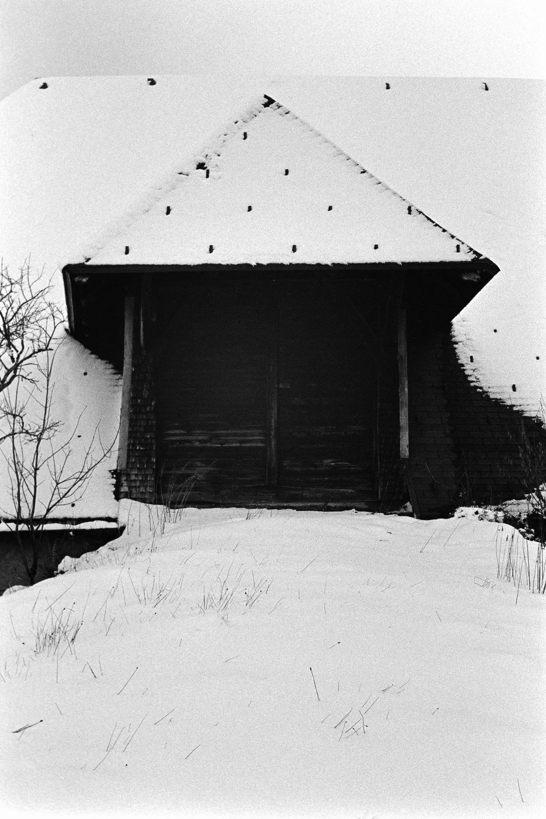 An old wooden barn with a steep roof covered in snow, situated on a snowy landscape with bare trees nearby.