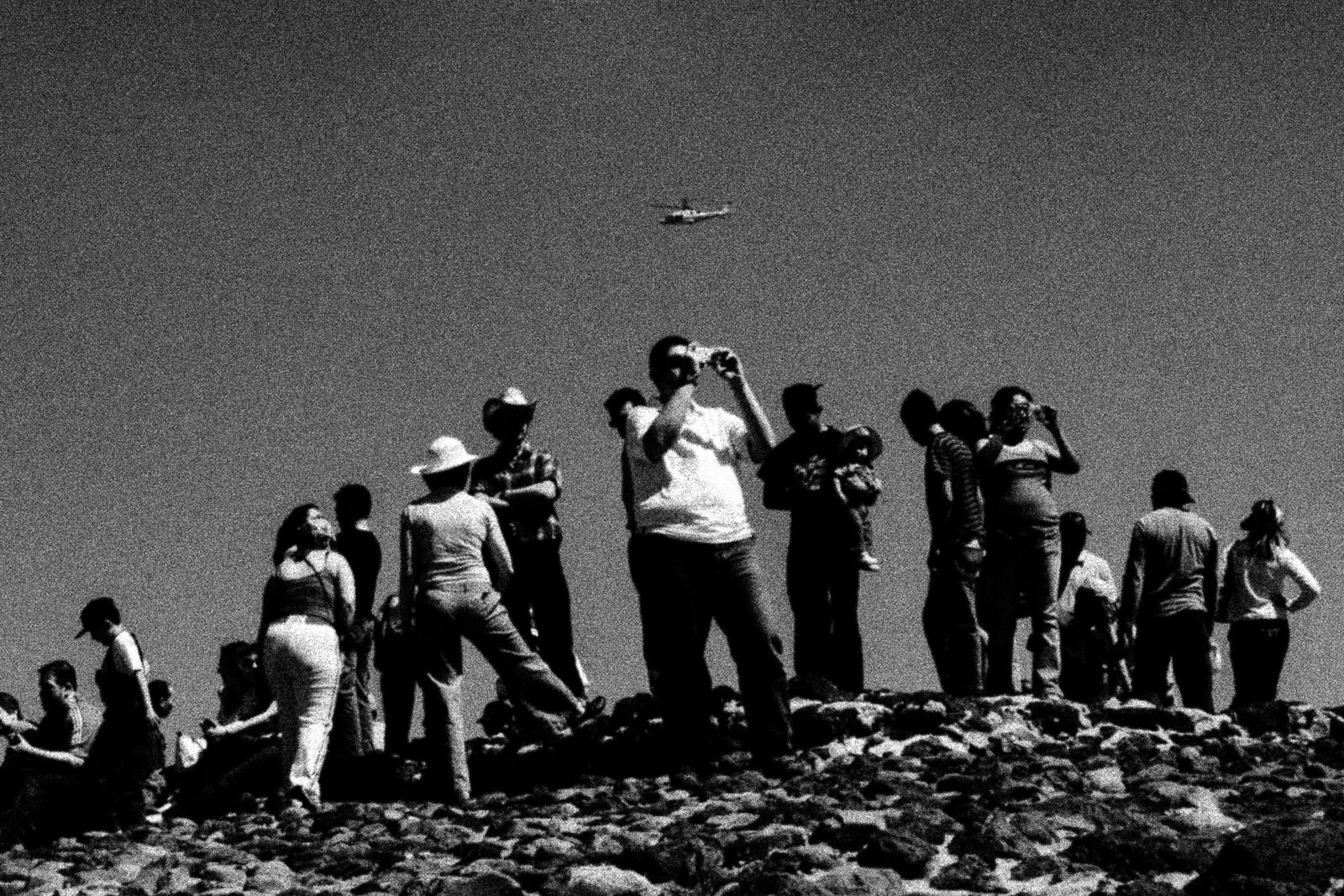 Group of people standing on rocky terrain, some holding cameras or phones, with an airplane flying overhead in the clear sky.
