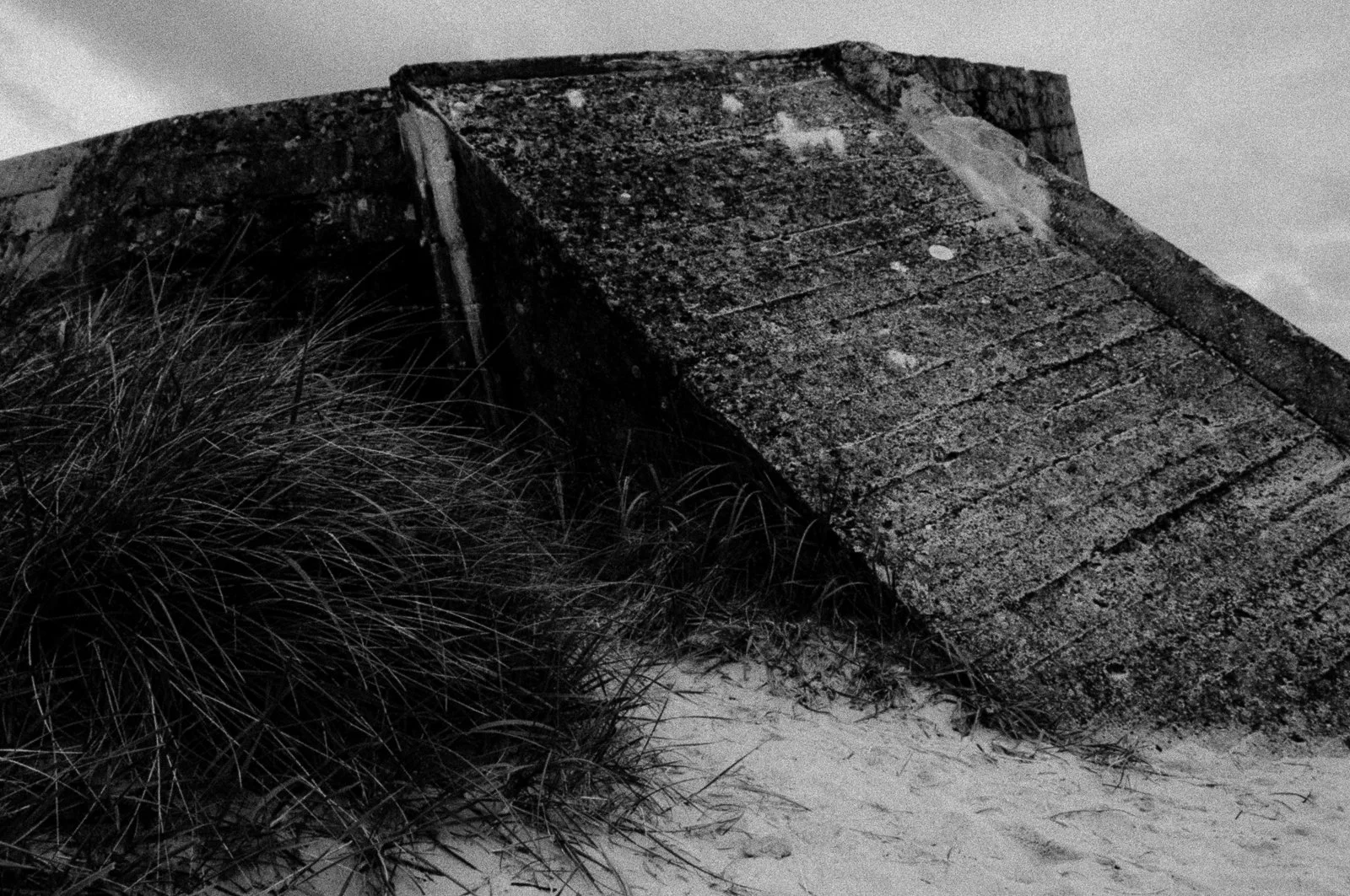 A black and white photograph of an old concrete bunker or fortification partially buried in sand, with tall grass growing nearby.