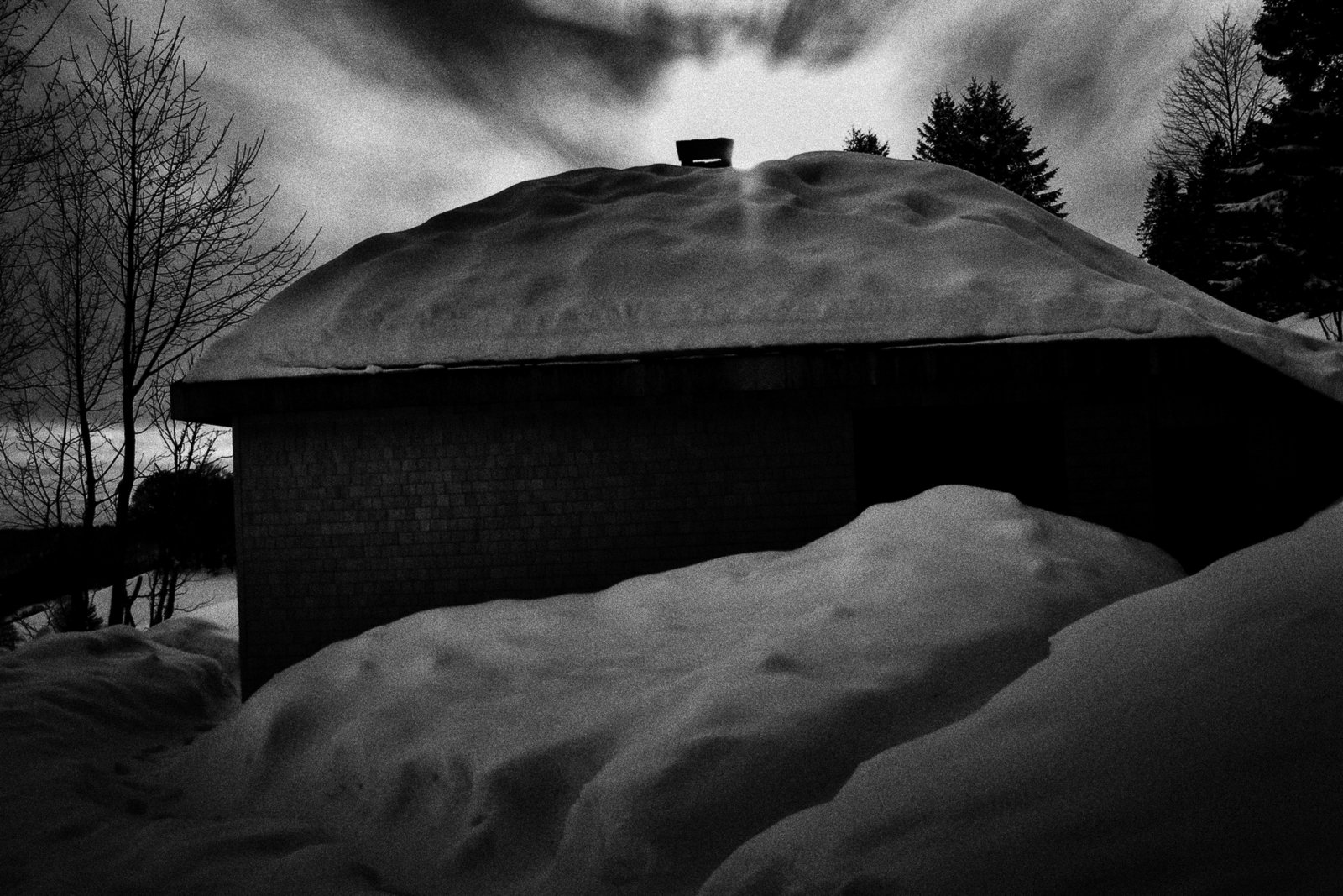 A house with a snow-covered roof and a large pile of snow in the foreground, trees without leaves, and a cloudy sky at dusk or dawn.