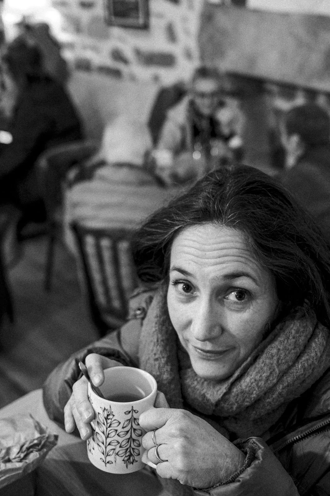 A woman with wavy hair looking up while holding a mug with a leaf pattern, seated at a table in a cozy cafe with brick walls and other patrons in the background.