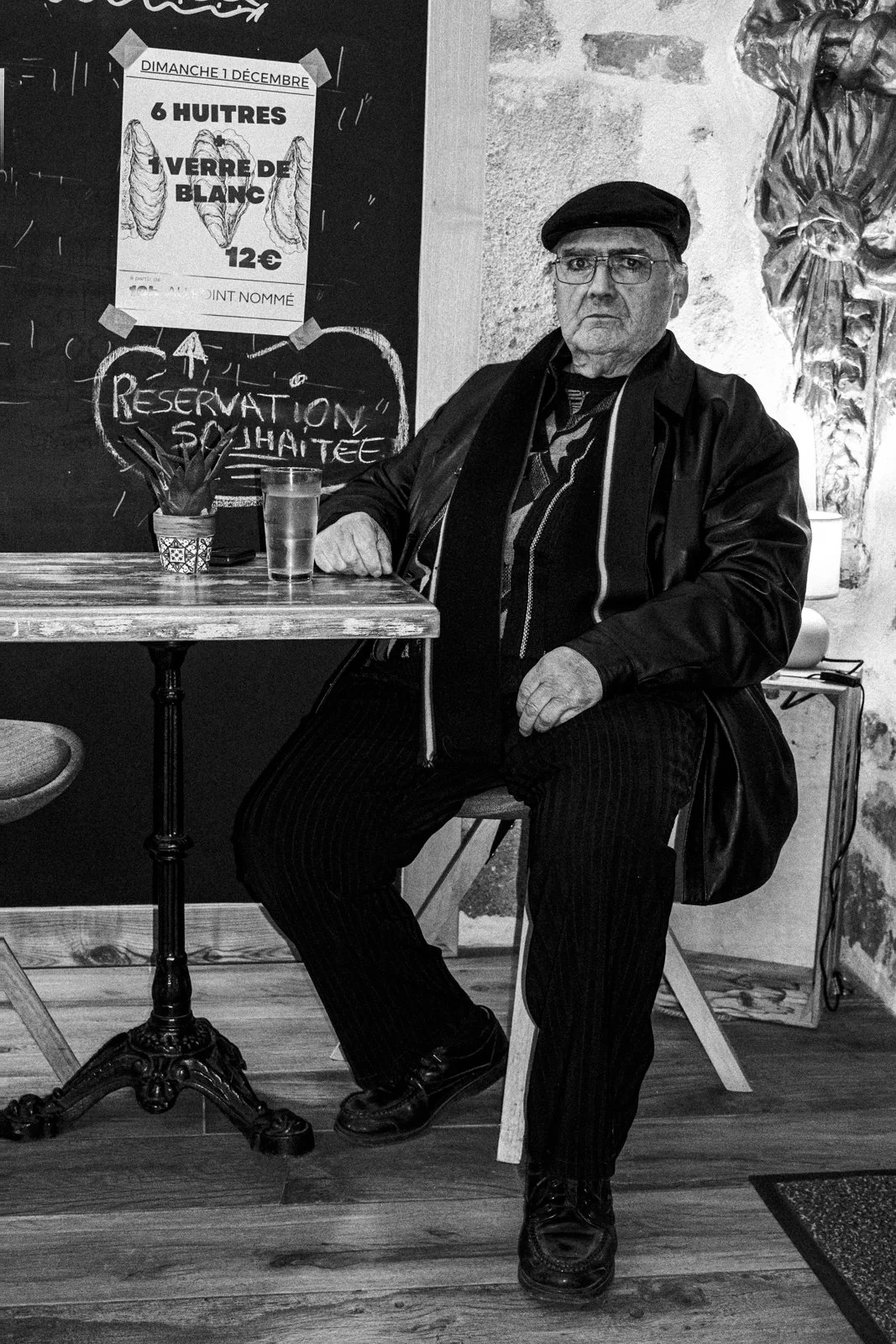 An elderly man sitting at a wooden table in a cafe, wearing a hat and glasses, with a serious expression. The table has a glass of water and a small plant. Behind him, there are signs and chalkboard art on the wall, including a handwritten message th