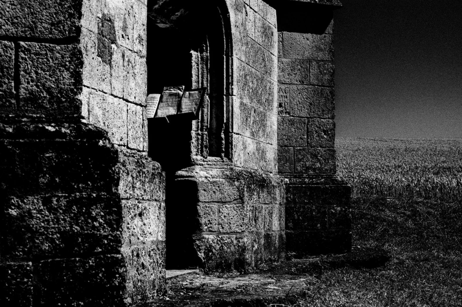 Black and white photo of an old stone building with an arched window, featuring two books and papers inside. A vast field is visible in the background.