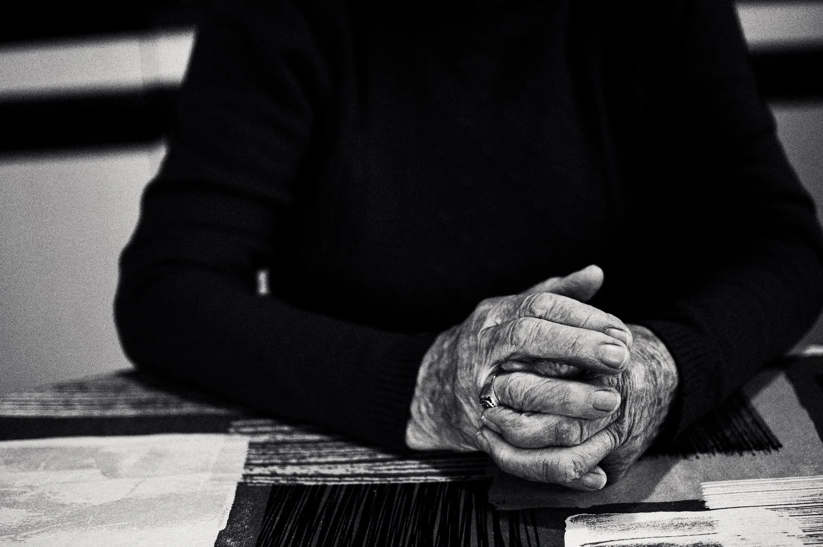 Close-up of elderly person's clasped hands resting on a table, wearing a dark sweater, in black and white.