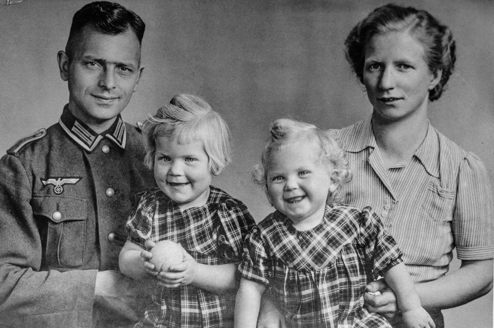 Black and white photo of a man dressed in a Nazi military uniform with two young girls, all smiling, against a plain background.