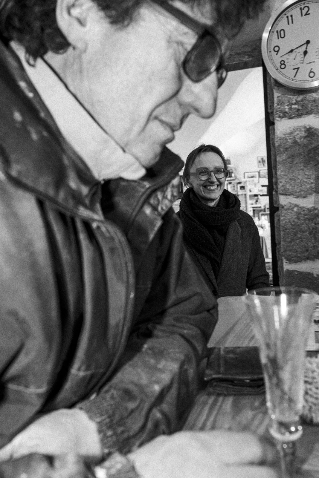 A man wearing glasses and a leather jacket is in the foreground, with a woman in the background smiling warmly. There is a clock on the wall showing the time as 8:43, and glasses and a wallet are on the table.