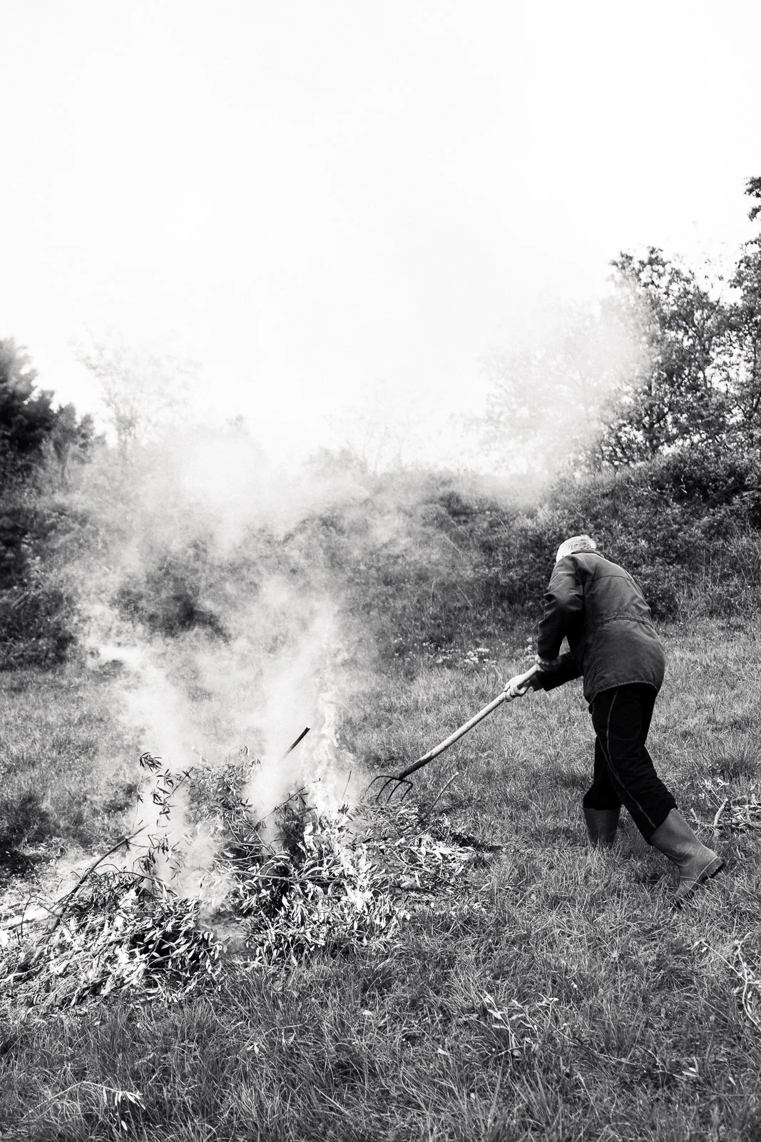 A person wearing a coat and boots using a rake to clear a small fire in a grassy field surrounded by trees.
