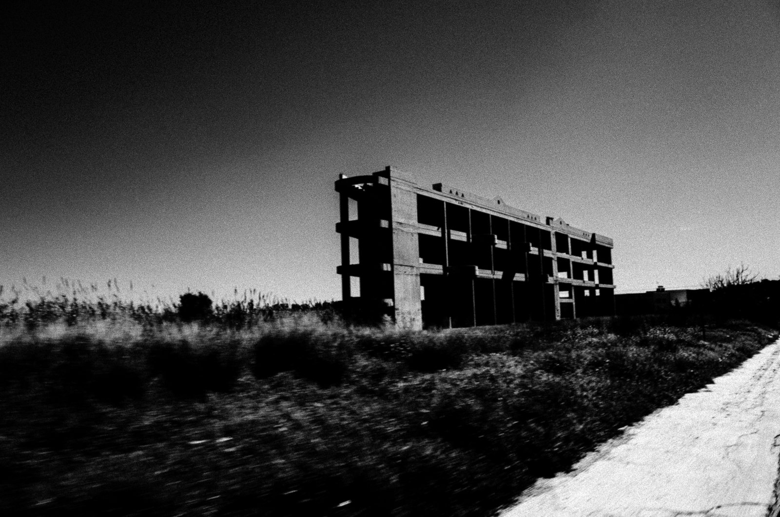 Black and white photo of an abandoned concrete building on a barren landscape with a dirt road in the foreground.