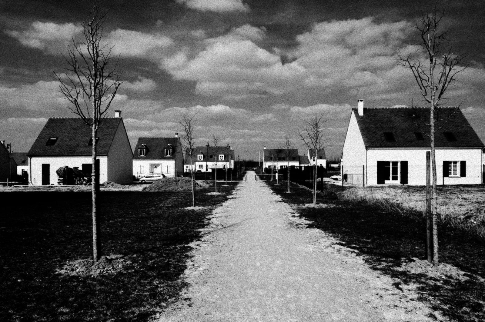 Black and white photo of a suburban neighborhood with houses, leafless trees, a dirt road, and a cloudy sky.