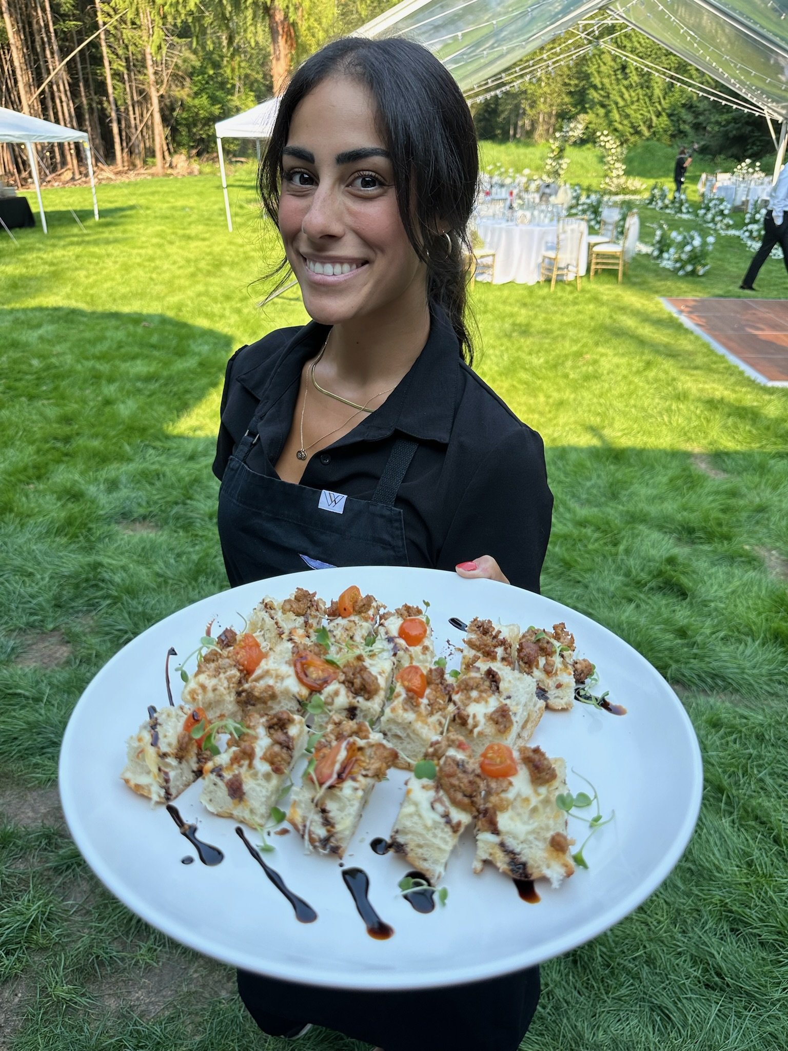 Smiling woman holding a white plate with appetizers, outdoor event with tables and canopy in the background.