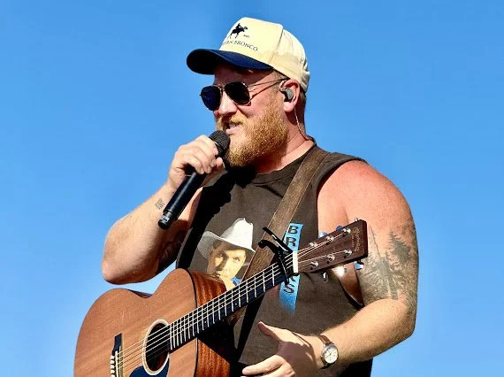 Man with sunglasses, a beard, and a cap, playing an acoustic guitar and singing into a microphone outdoors against a blue sky.
