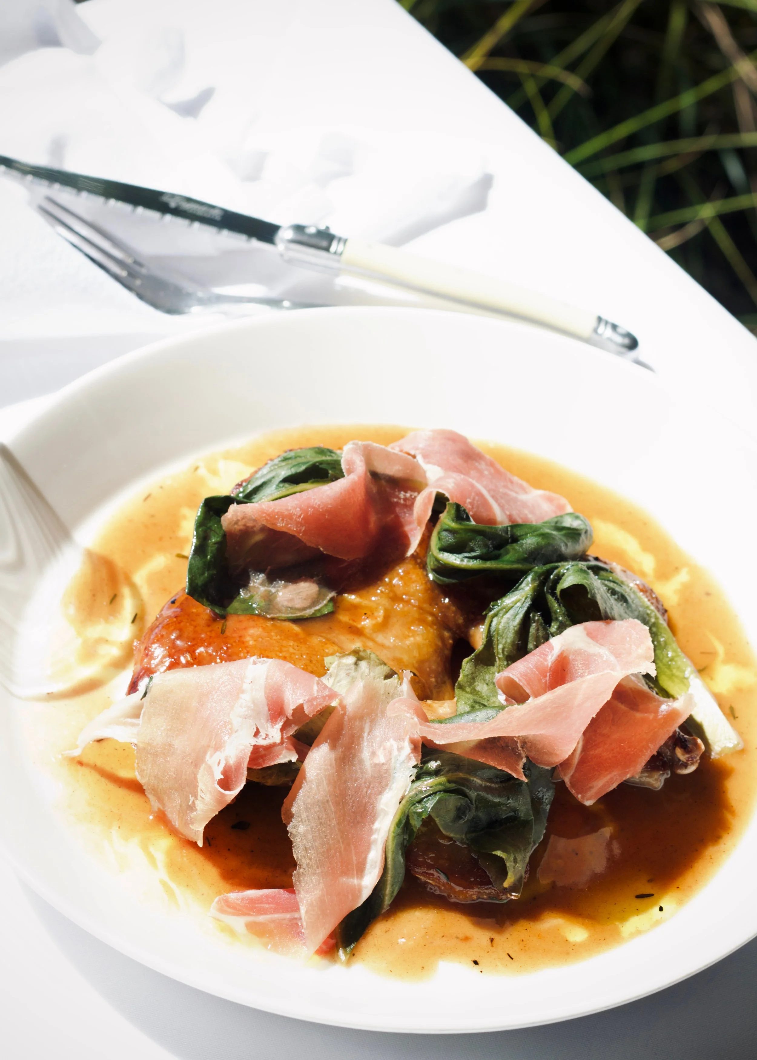 A gourmet dish featuring slices of prosciutto and greens on top of a cooked piece of poultry in a brown sauce, served on a white plate with a fork and knife nearby.
