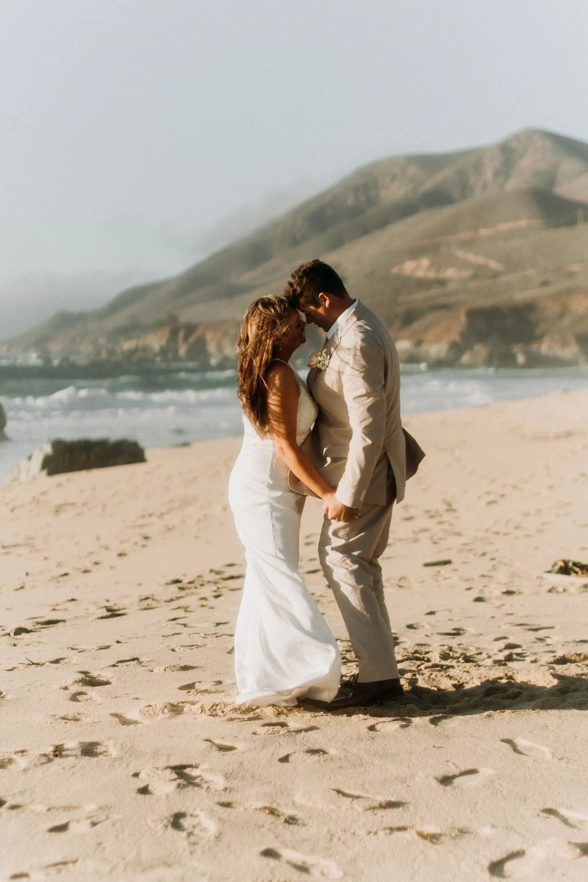 April and Trey embracing on the beach with the cliffs in the background during their Big Sur elopement