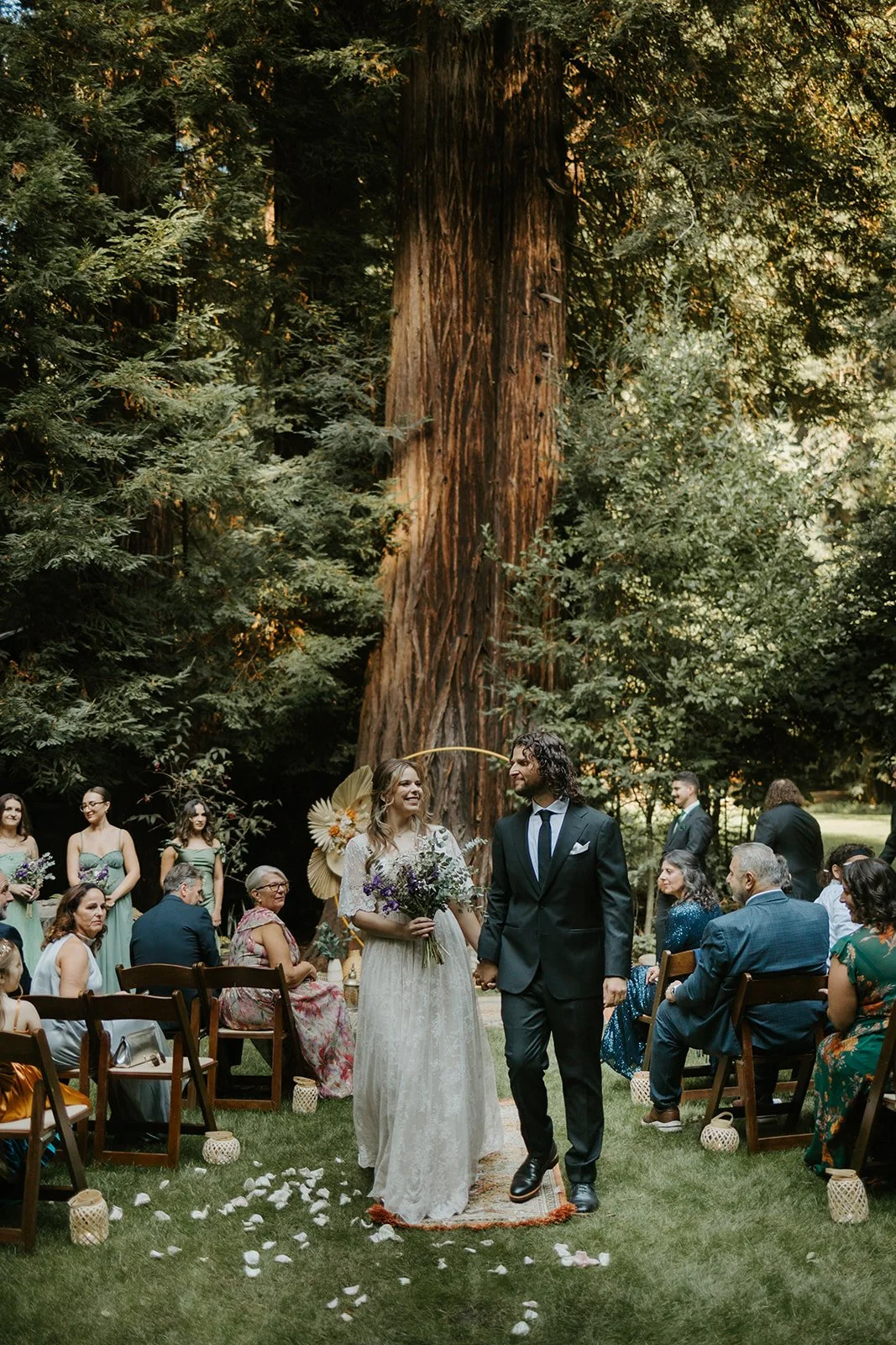 A bride and groom share a kiss outdoors on their wedding day, with the bride holding a bouquet of pink and white flowers and wearing a lace wedding dress, while the groom is dressed in a gray suit.