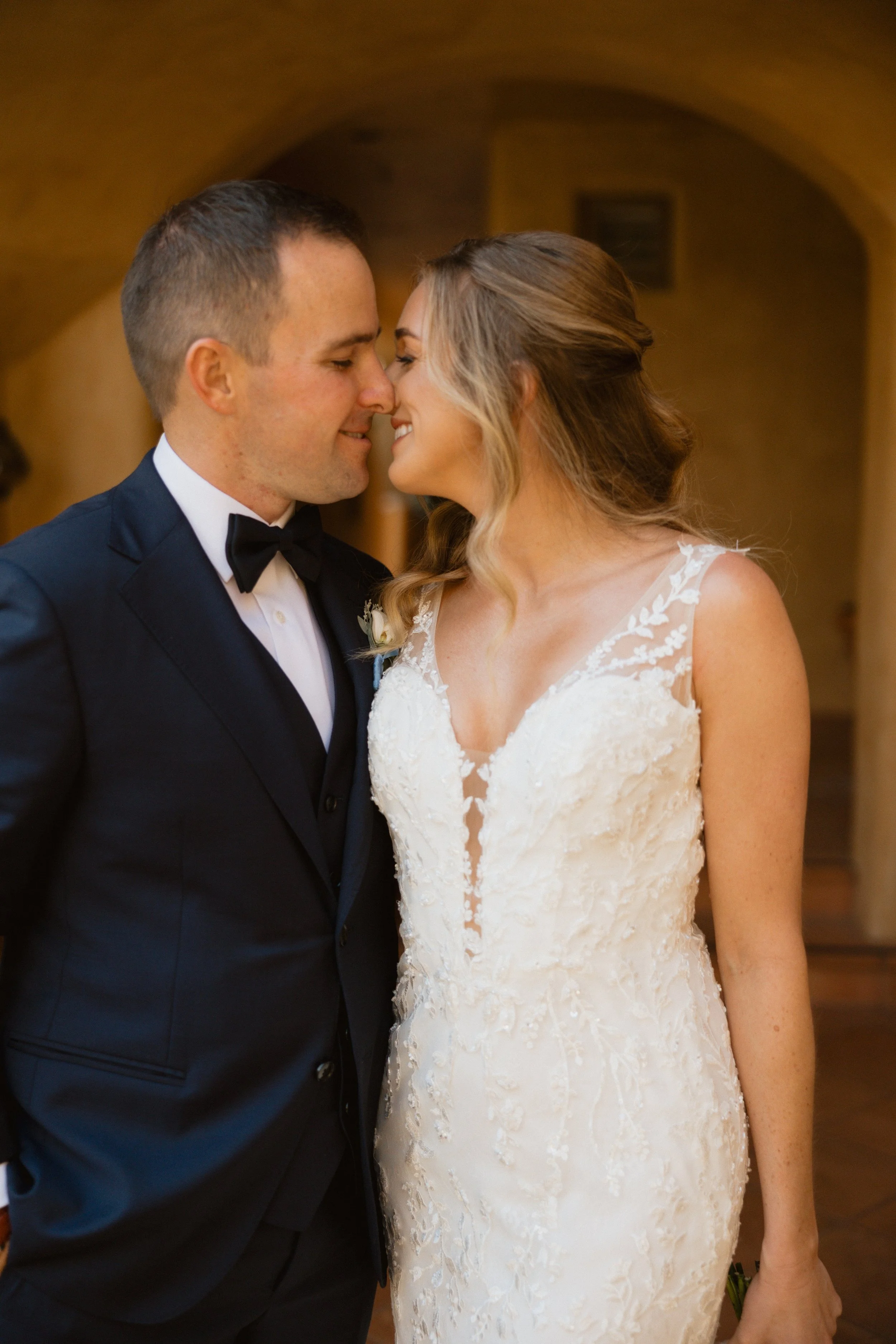 A bride and groom share a tender moment, about to kiss, with their noses touching indoors in a warm, softly lit setting.