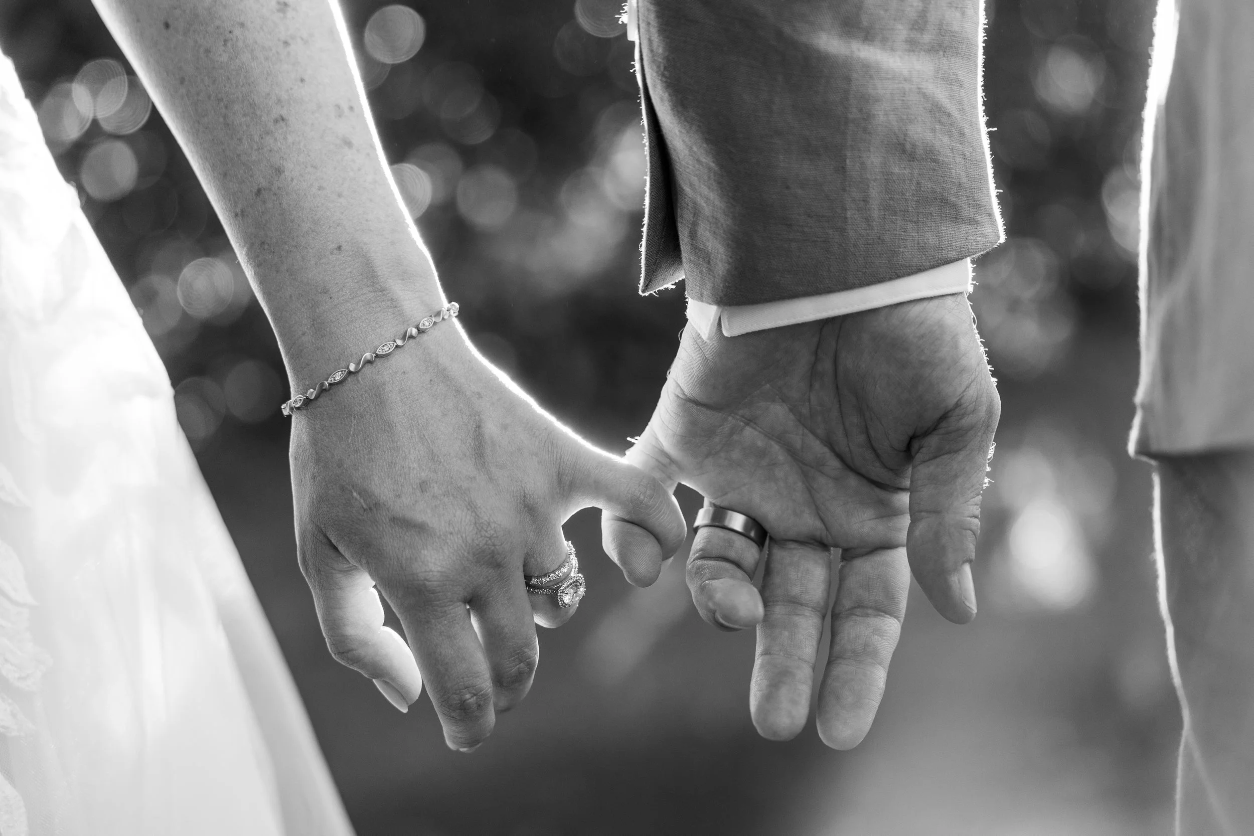 Black and white close-up of a couple holding hands during their wedding, with wedding rings visible on their fingers.