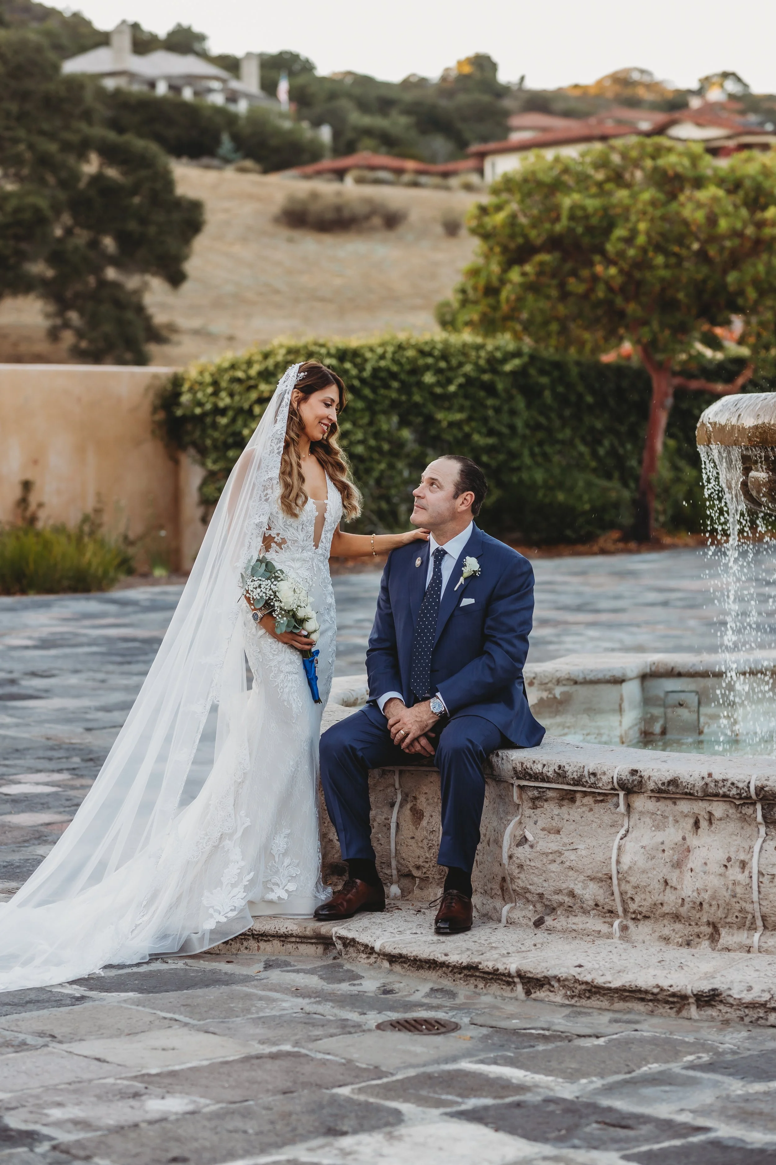 A bride in a white wedding gown with a veil and bouquet stands next to a groom in a navy suit sitting on a stone ledge by a fountain in an outdoor setting.