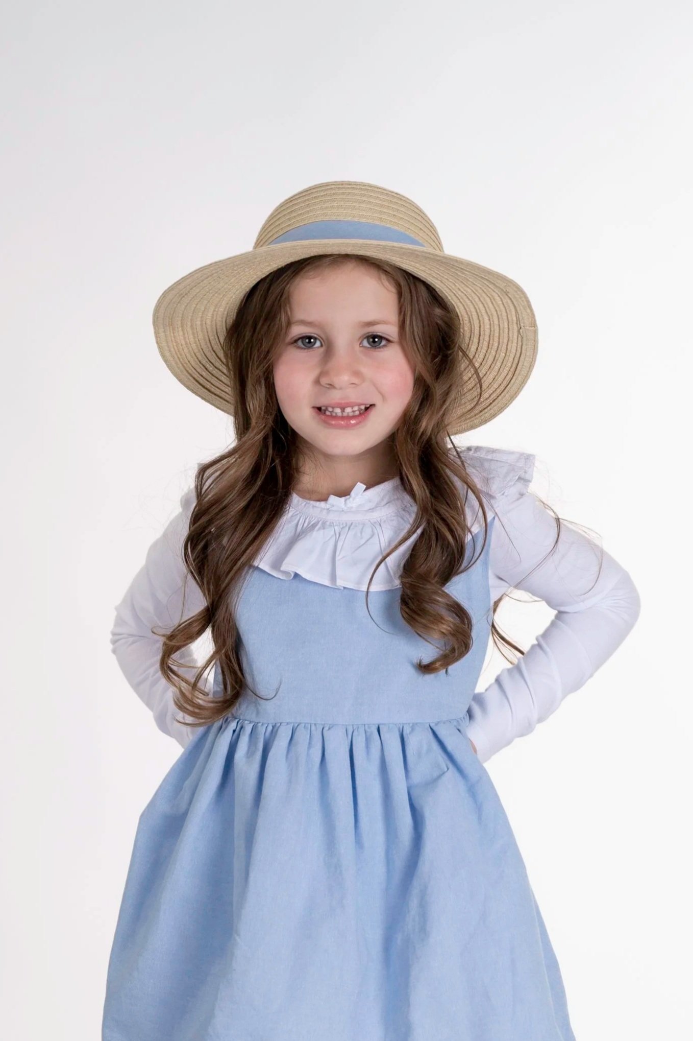 A young girl with long wavy hair wearing a white long-sleeve top, a light blue dress, white tights, white shoes, and a straw hat with a blue band standing on a wooden block against a white background.