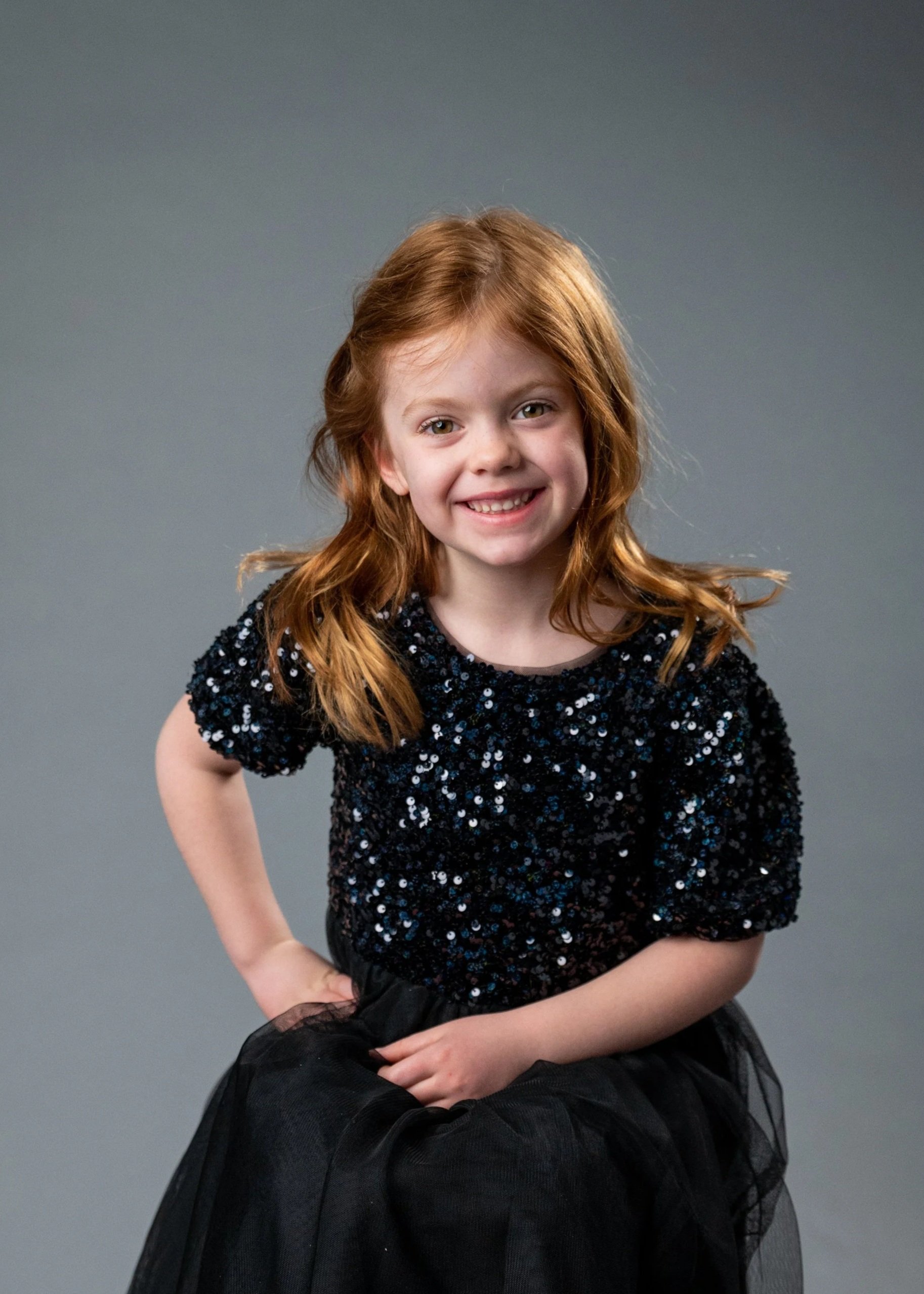 A young girl with red hair, smiling, sitting on a chair against a gray background. She is wearing a black, sparkly top, a black tulle skirt, white tights, and pink shoes with flower decorations.