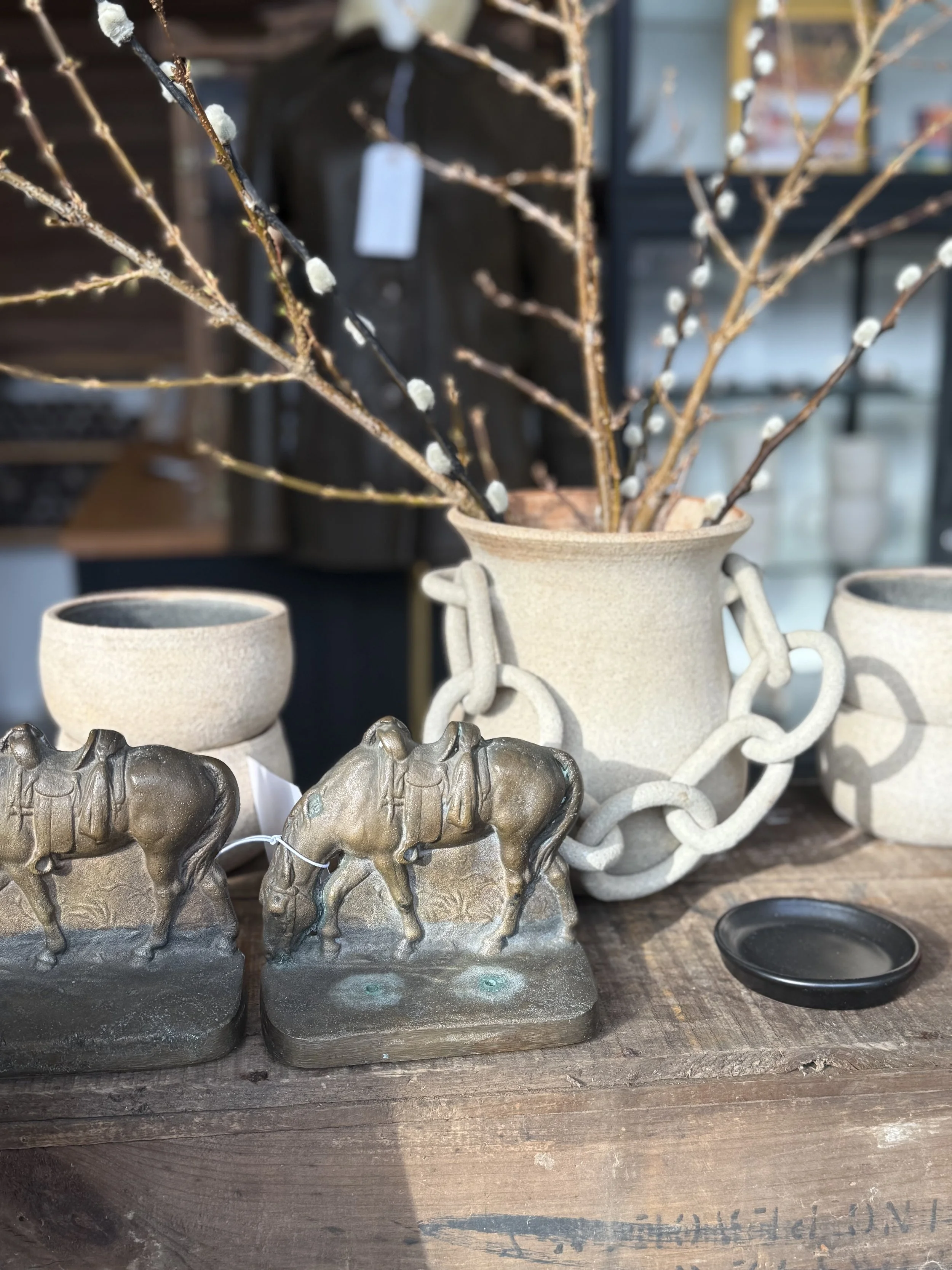 Decorative display featuring two horse figurines with saddles, beige ceramic pots, a black dish, and branches with white buds arranged in a large textured vase on a wooden surface.