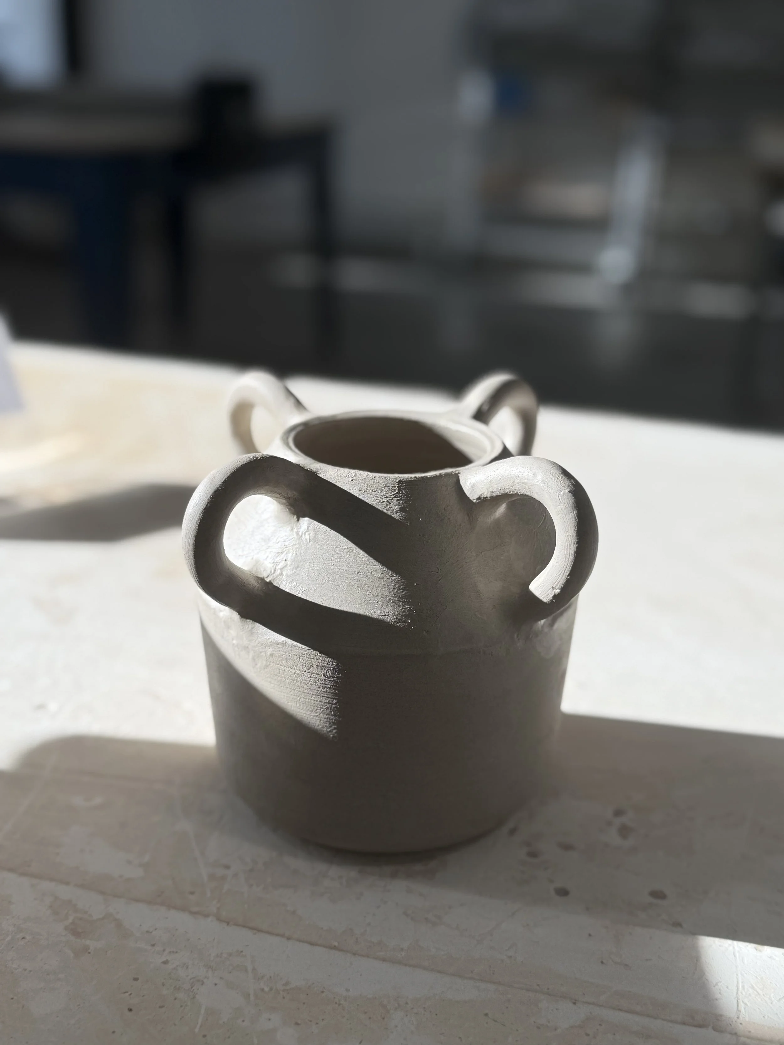 A ceramic pitcher with two small handles on a white surface, illuminated by sunlight, with a blurred background.