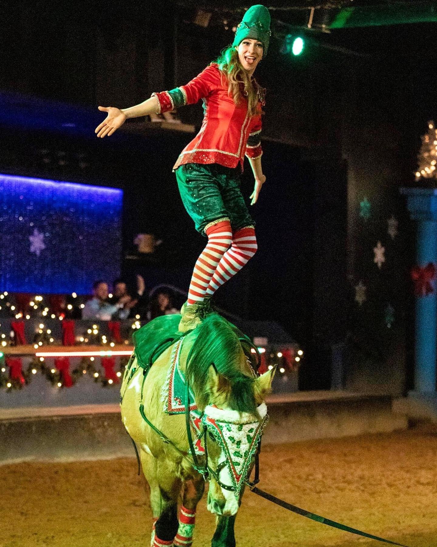 A female performer balancing on a horse during a Christmas-themed circus act. She is dressed in festive red and green clothing with striped stockings, smiling, and extending her arms.