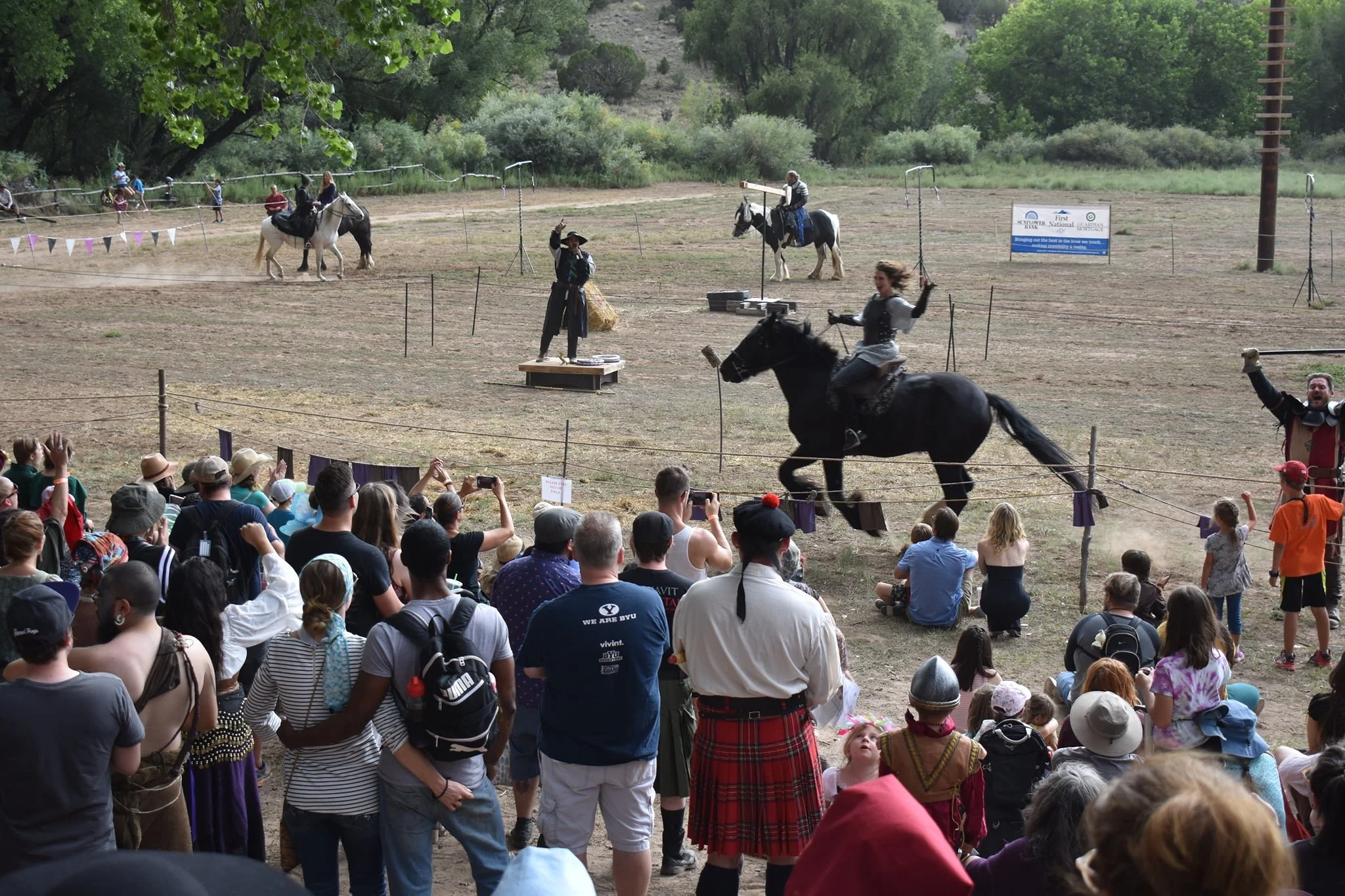 A medieval-themed reenactment featuring knights on horseback in armor, engaging in a staged combat or duel, with an audience of people in modern casual and historical costumes watching and taking photos outdoors in a field.